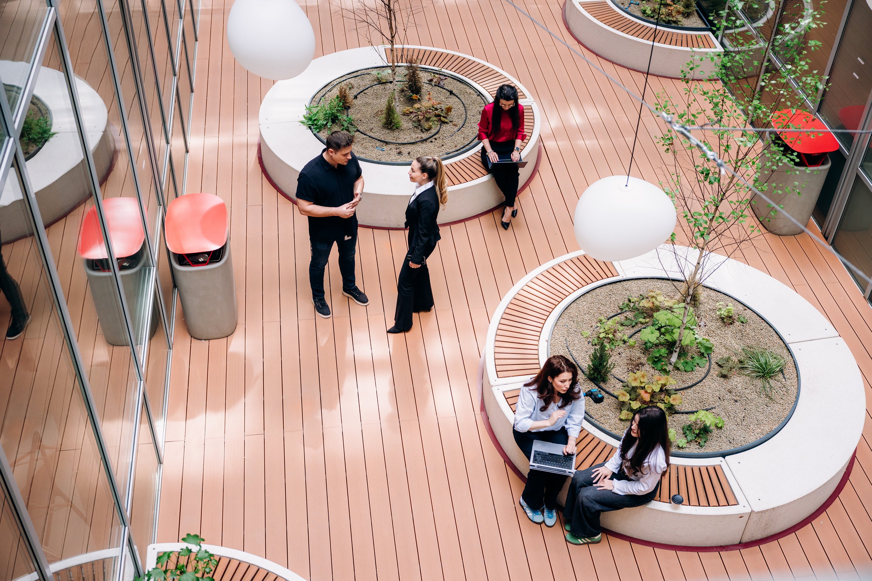 Diverse professionals collaborate and network within a modern, sunlit architectural office courtyard featuring circular seating and planters.