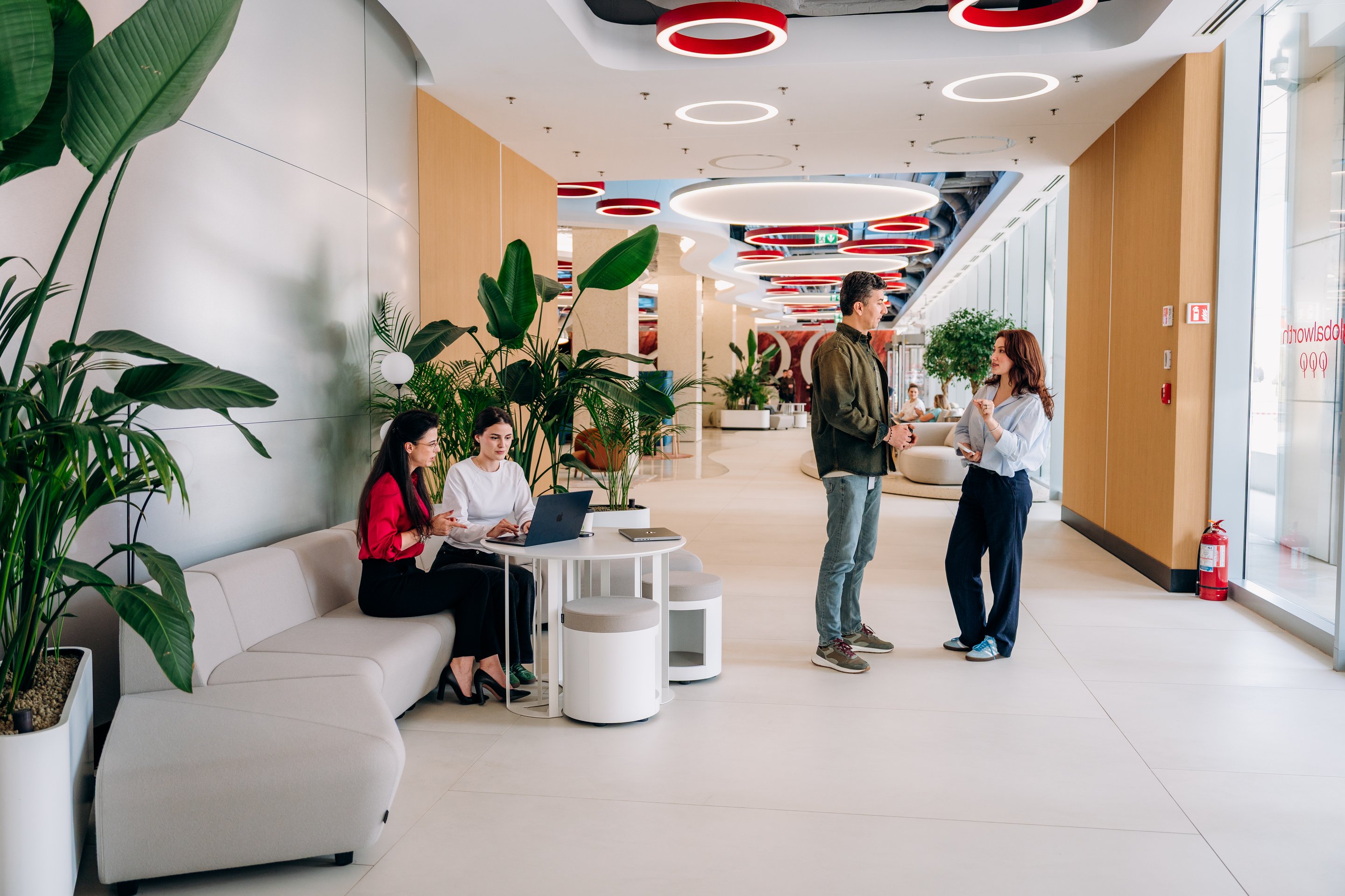 Diverse professionals engage in productive discussion and laptop-based work within a modern, plant-filled corporate office lounge.