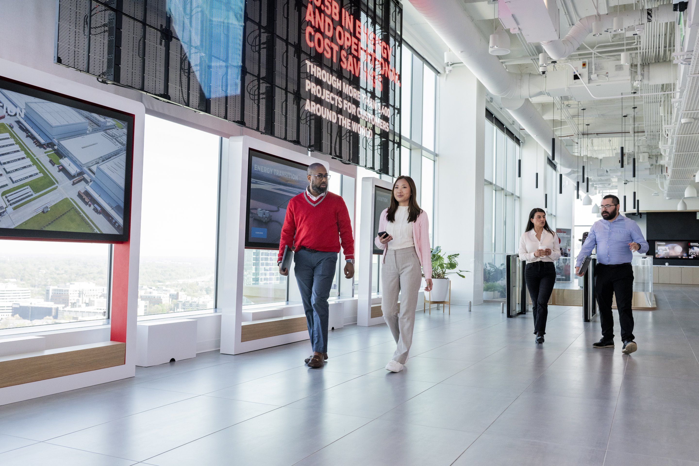 Diverse corporate team members walk and discuss business in a modern, light-filled office corridor with digital displays.