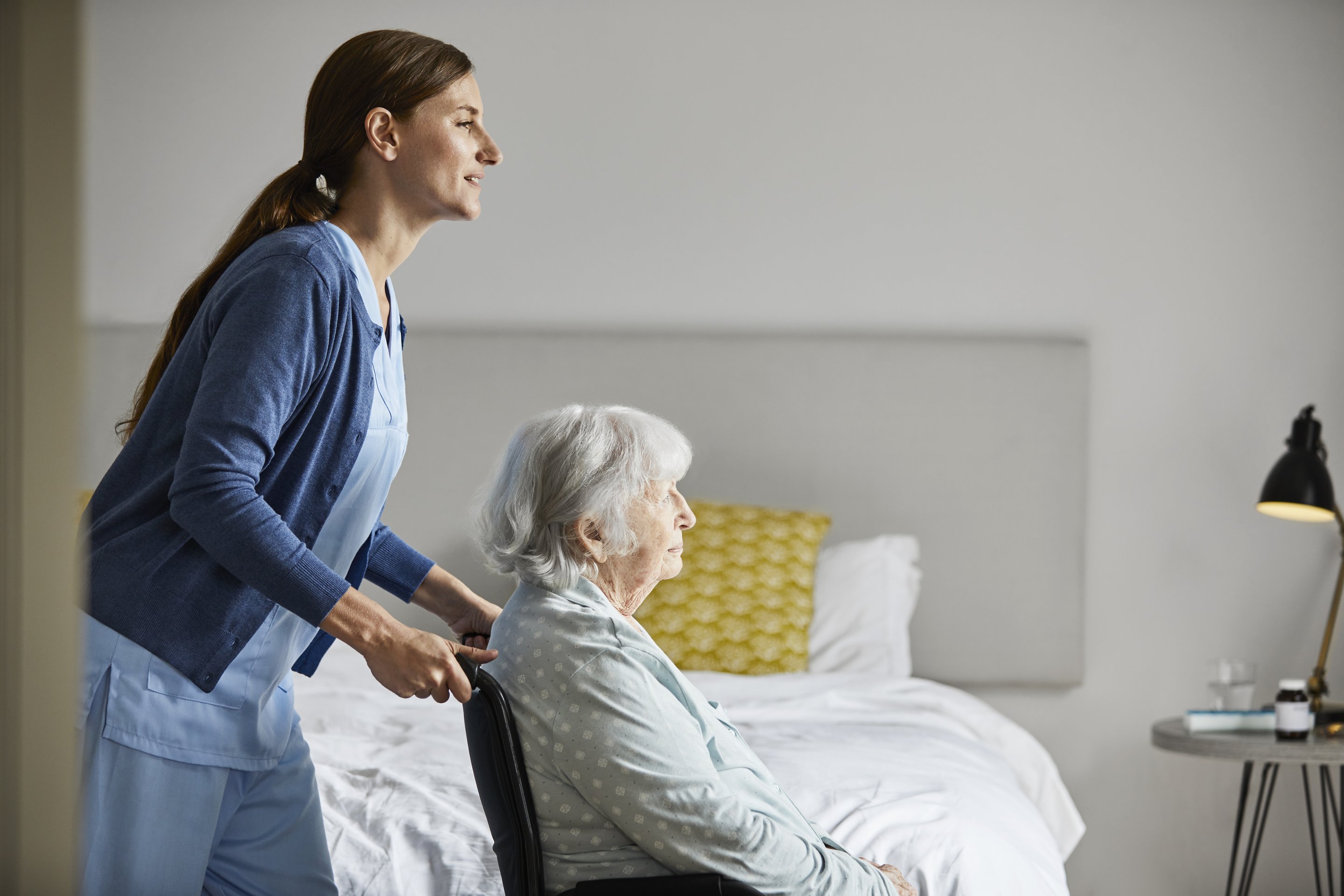 A professional caregiver assists an elderly woman in a wheelchair inside a comfortable, modern bedroom setting.
