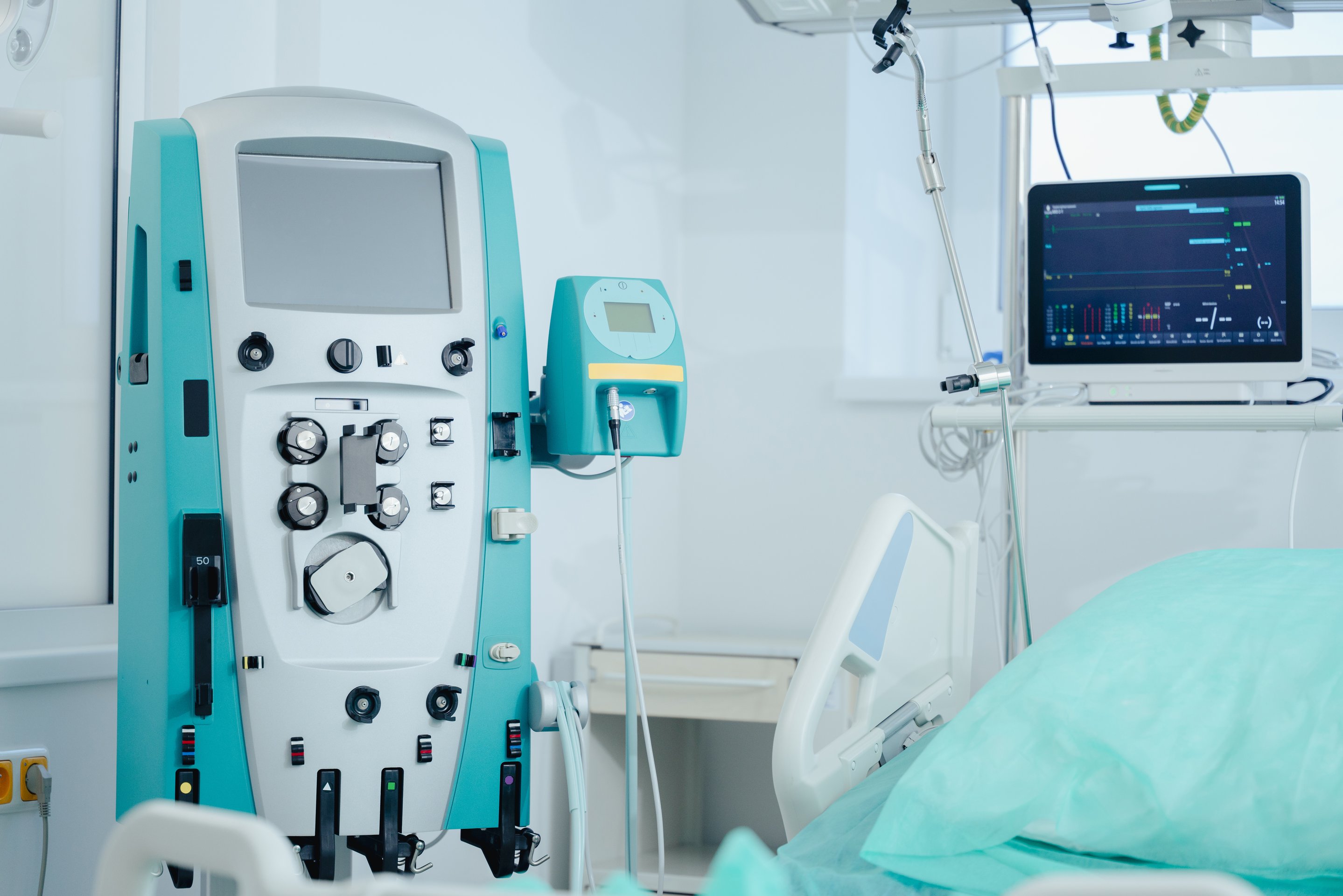 A clinical dialysis machine stands next to a hospital bed with a patient monitor displaying vital signs in a sterile medical treatment room.