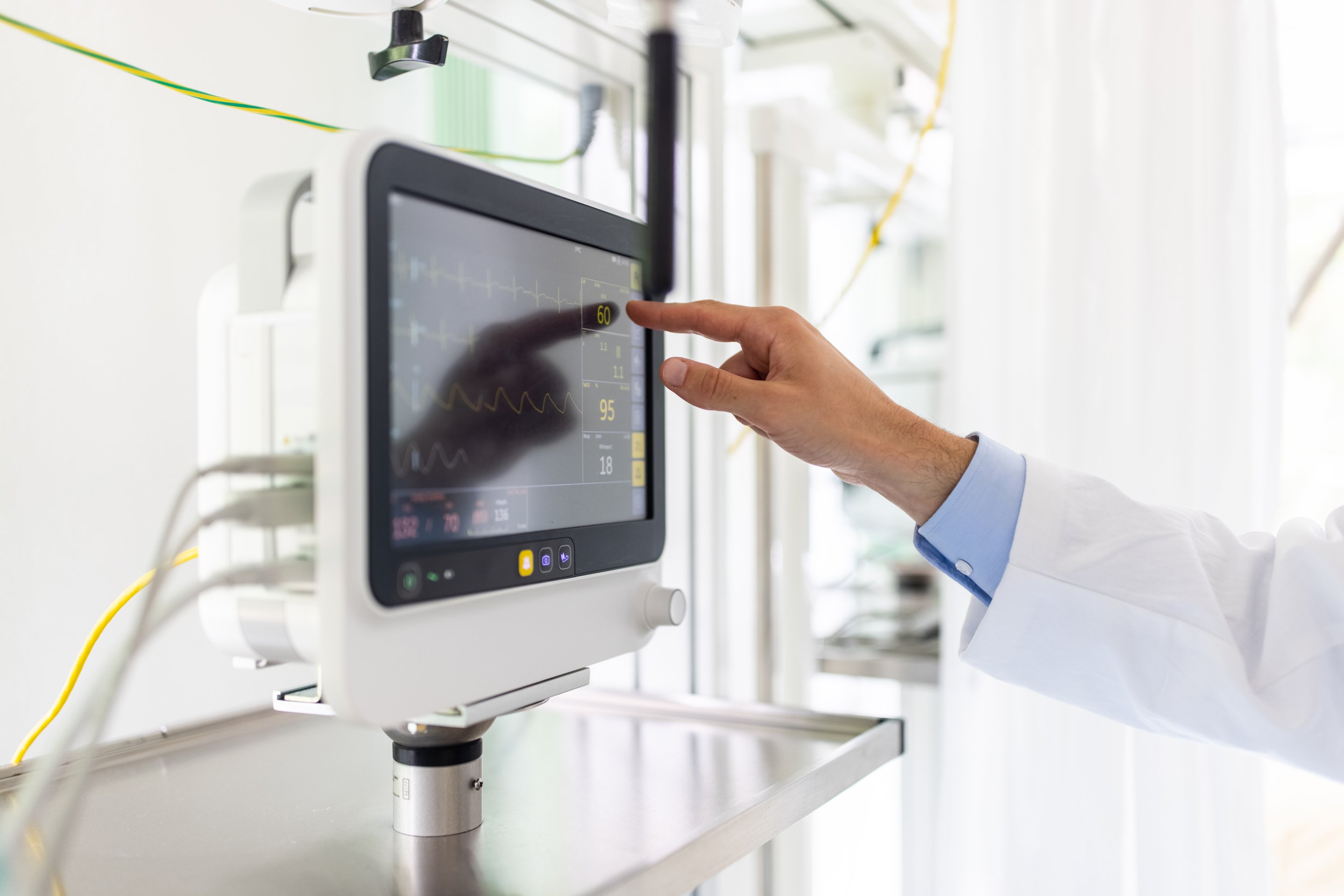 A medical professional in a white coat monitors vital sign readings on a digital hospital patient display screen.
