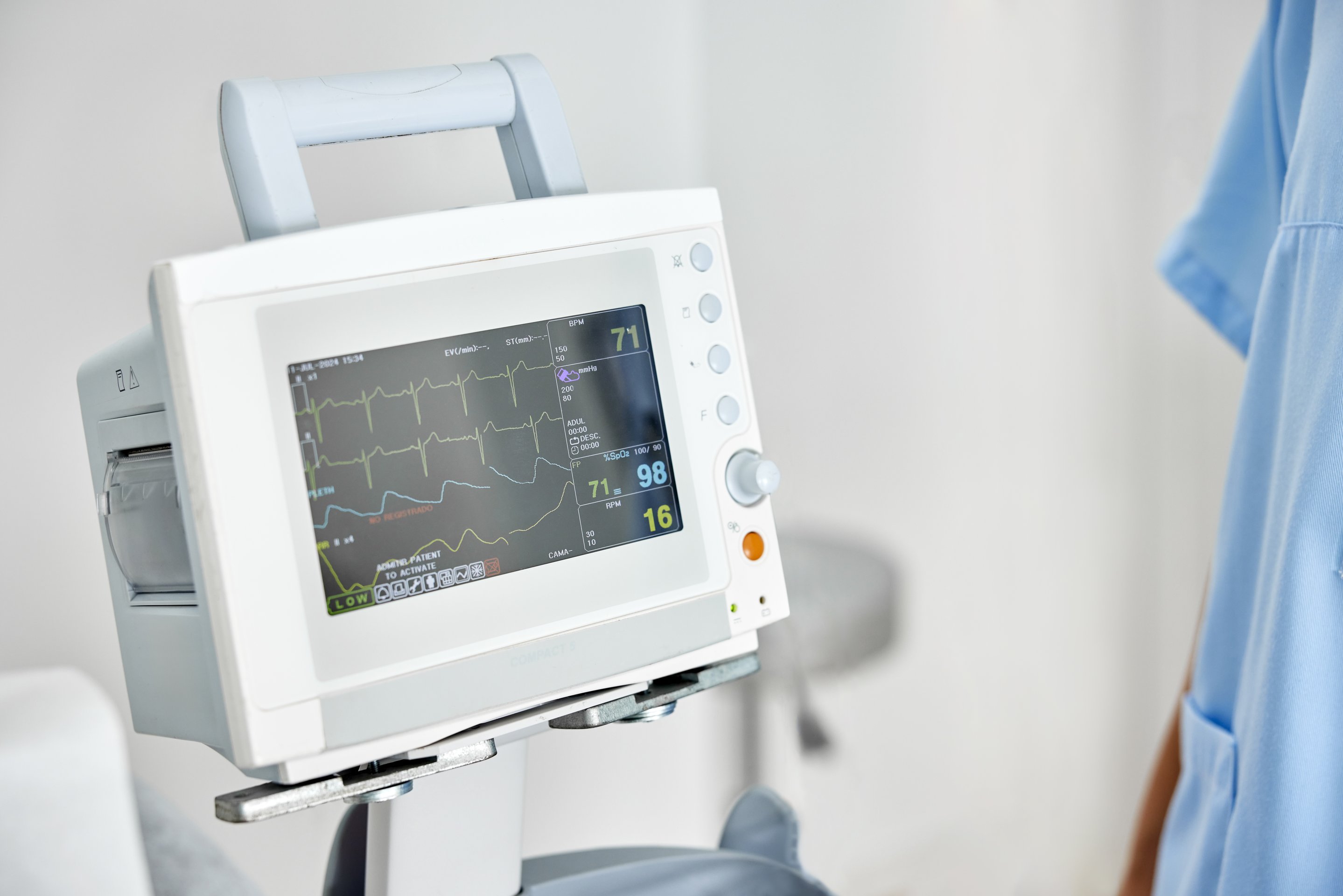 A close-up view of a medical patient monitor displaying real-time vital signs with a healthcare professional standing in the background.