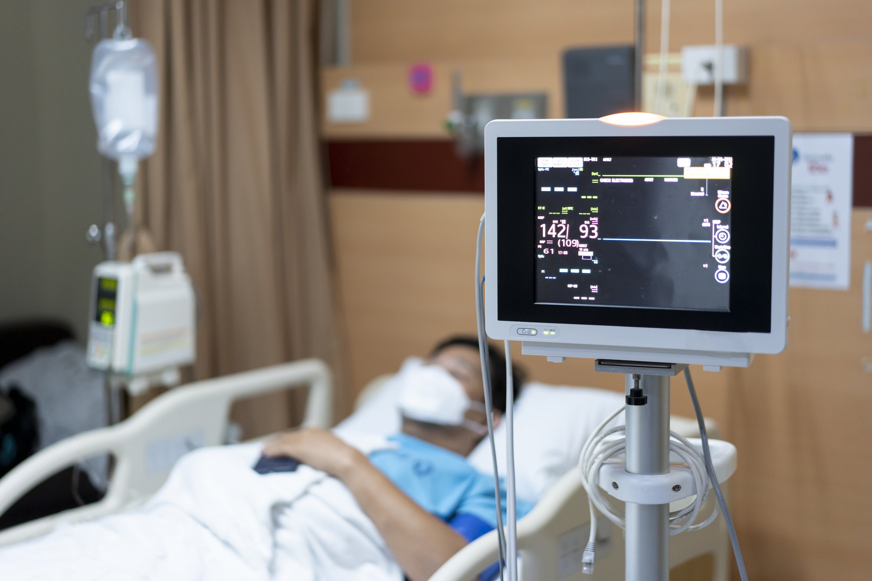 Digital bedside monitor displaying vital signs for a blurred patient resting in a hospital bed.
