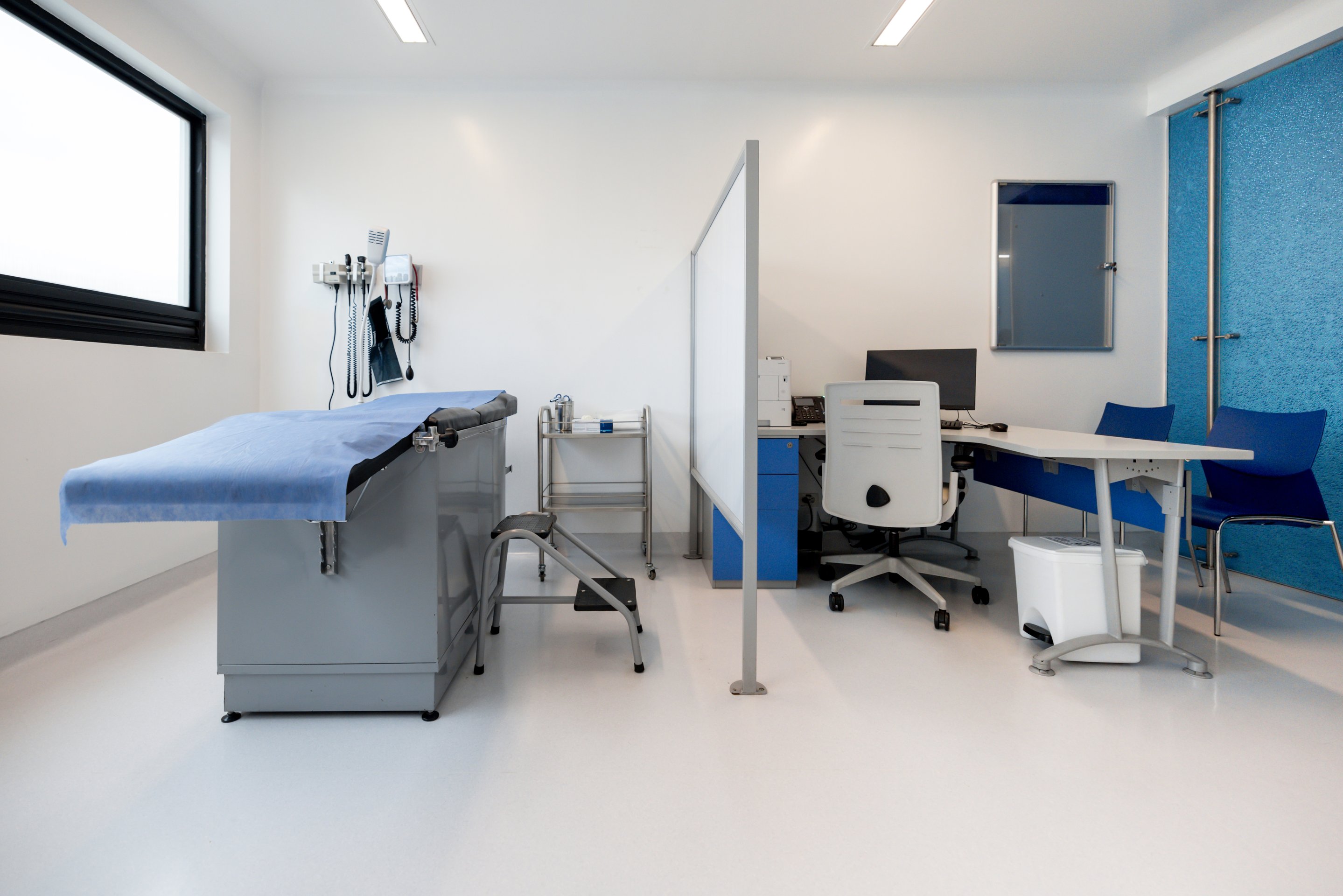 A clean, well-lit medical examination room featuring a patient bed, a divider screen, and a professional workspace area.