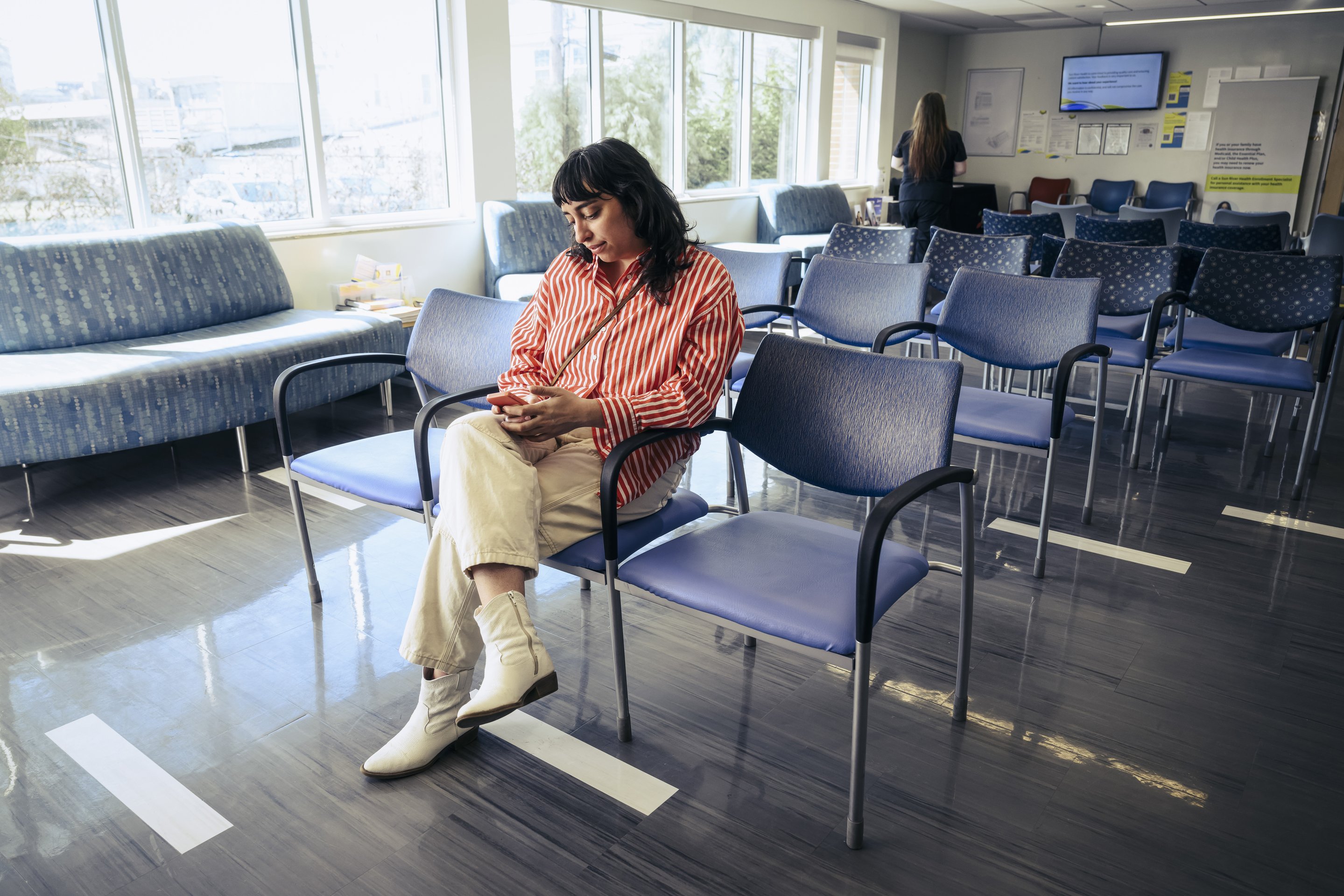 A woman in a striped shirt sits in a brightly lit office waiting area while looking at her smartphone.