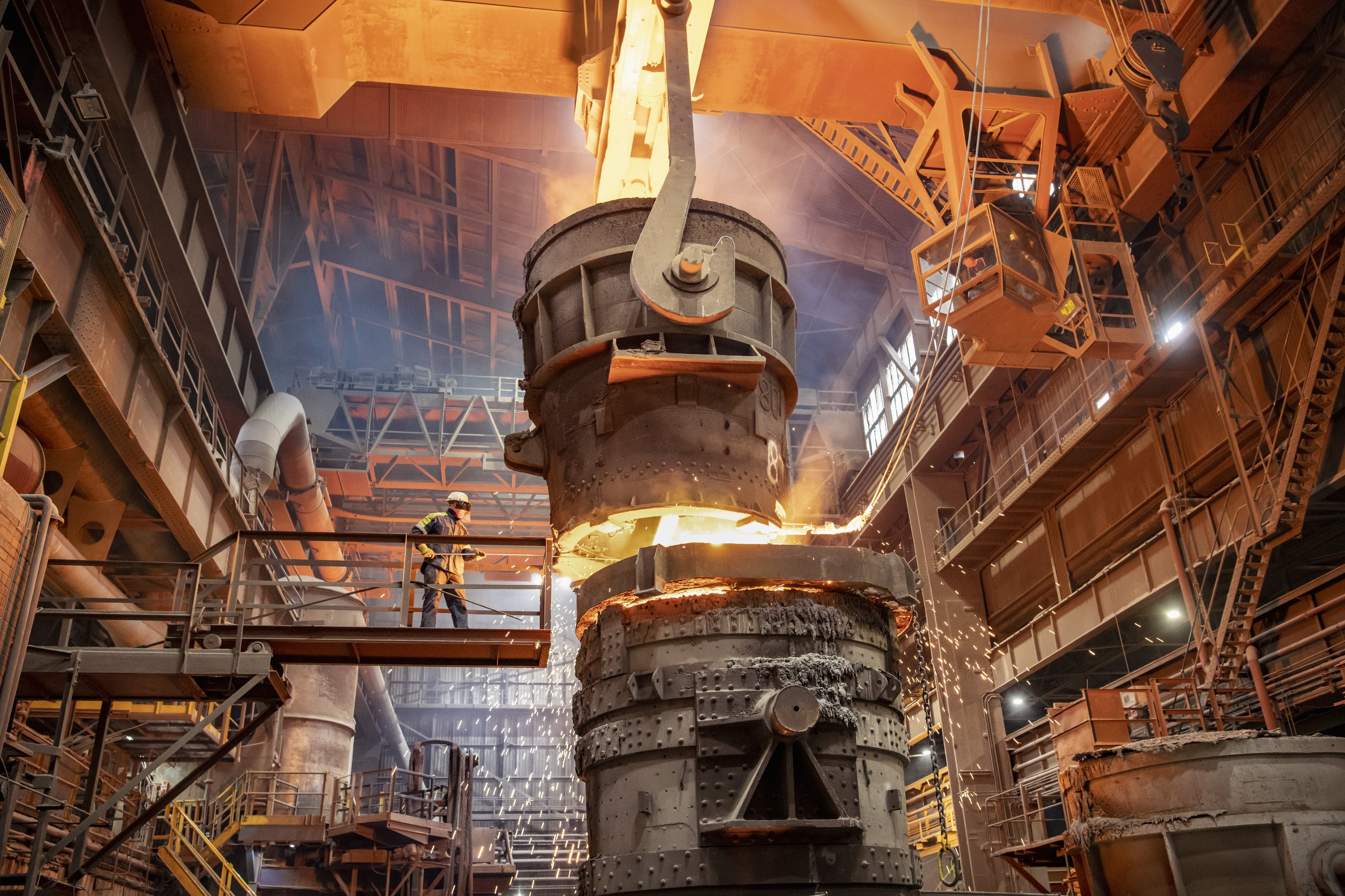 An industrial worker oversees the pouring of molten metal from a crane-suspended ladle into a secondary container at a steel manufacturing plant.