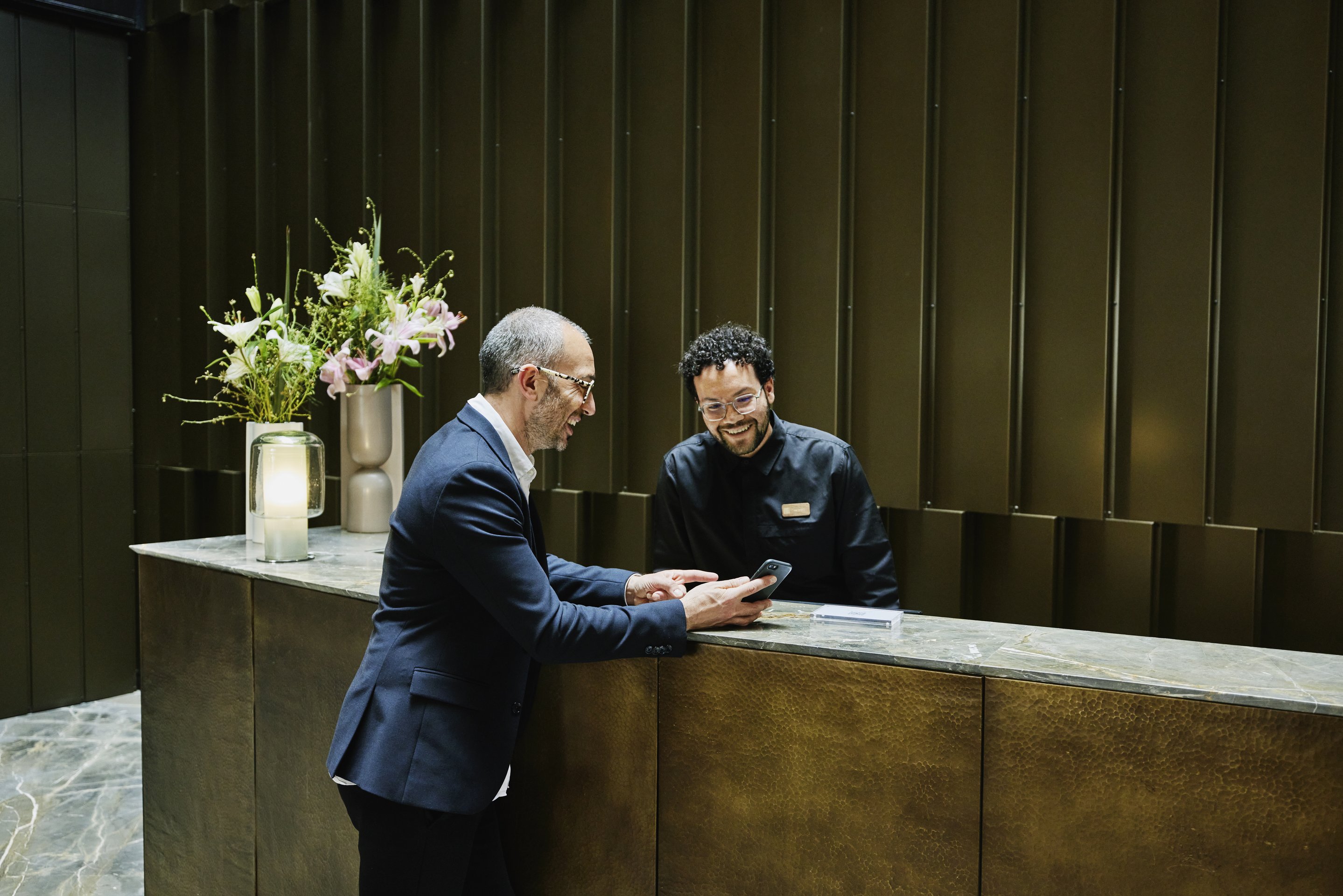 A professional concierge and businessman share a friendly interaction at a modern luxury hotel reception desk.