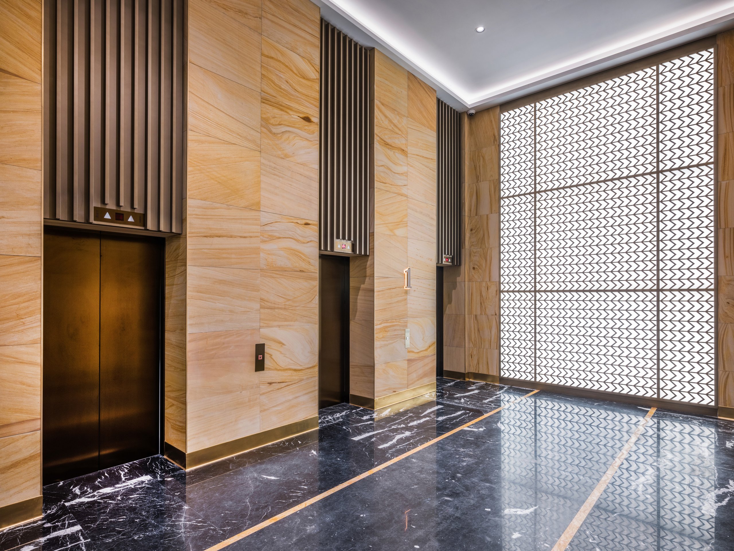 A contemporary corporate lobby featuring sleek bronze elevators, textured stone walls, and a large decorative backlit feature wall over polished marble floors.