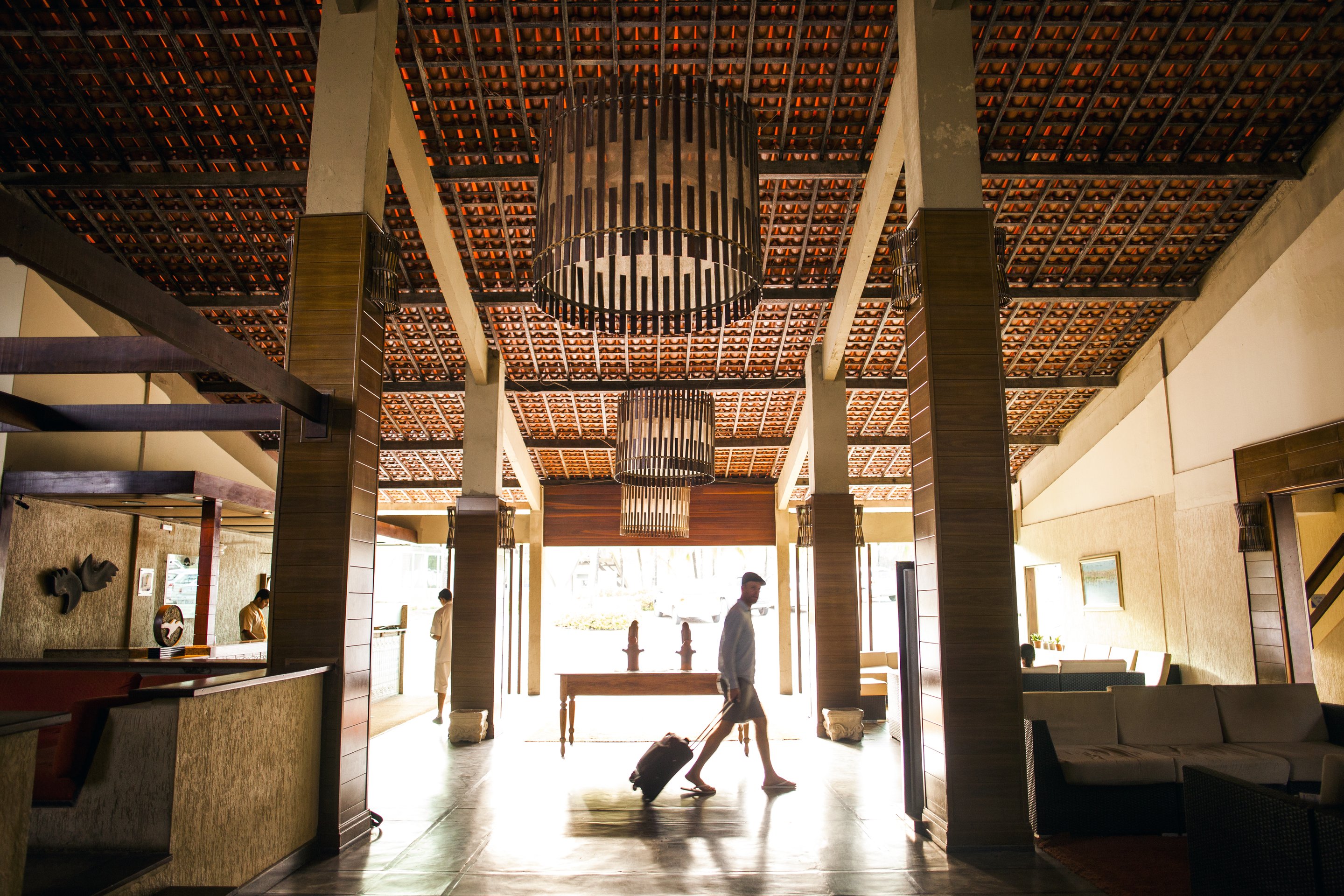 A man with luggage walks through a spacious, high-ceilinged hotel lobby with warm wood accents and contemporary design elements.