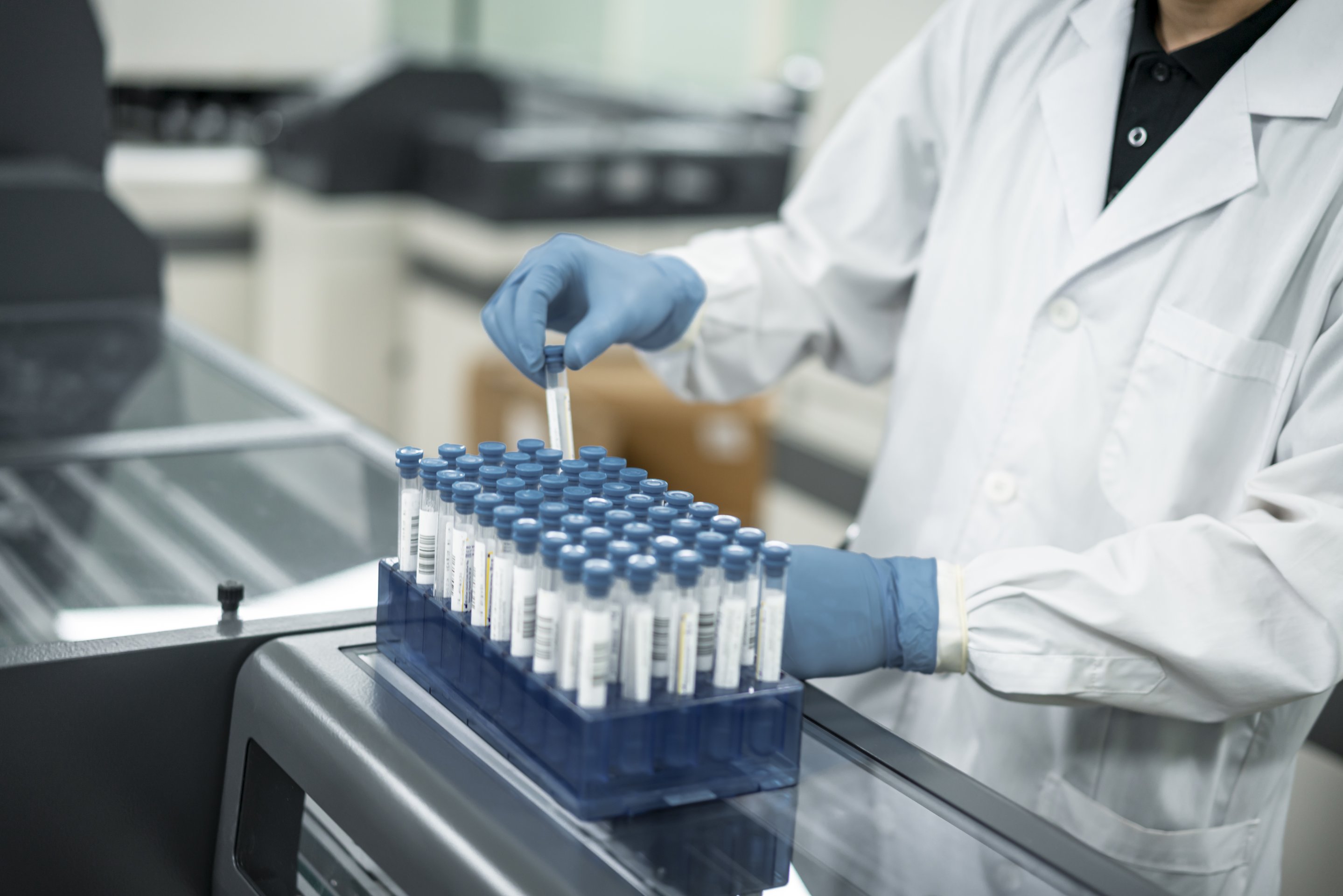 A technician in a lab coat and protective gloves processes a rack of test tubes in a modern clinical diagnostic laboratory.