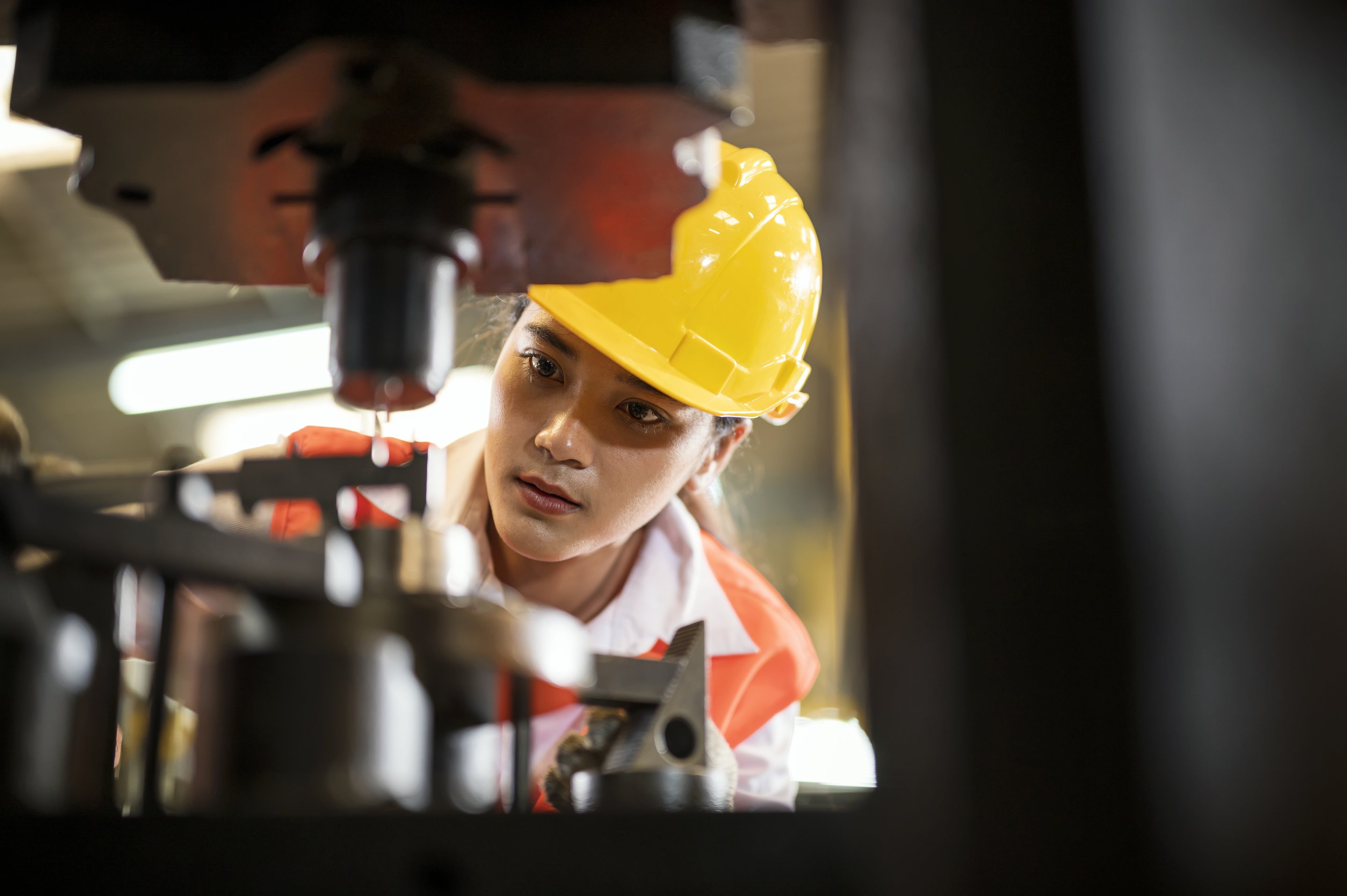 A focused female engineer in a yellow hard hat carefully monitors industrial machinery during production.