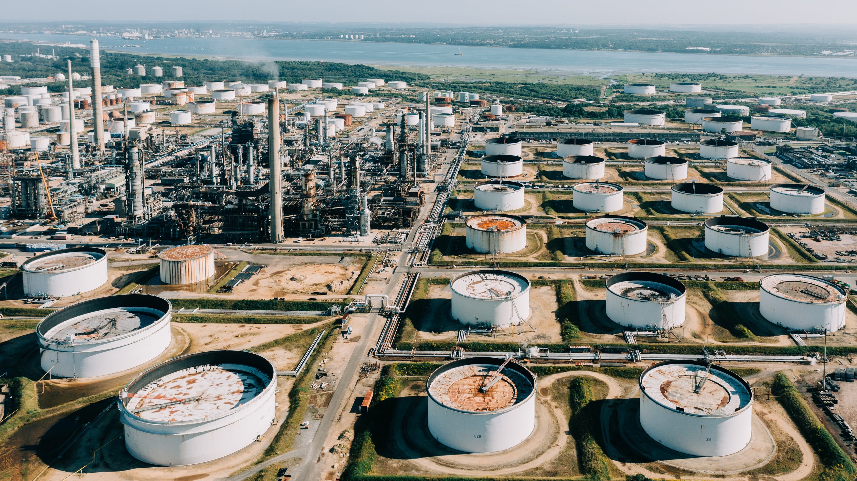 An aerial view shows an extensive oil refinery complex featuring large storage tanks and processing infrastructure near a coastal landscape.