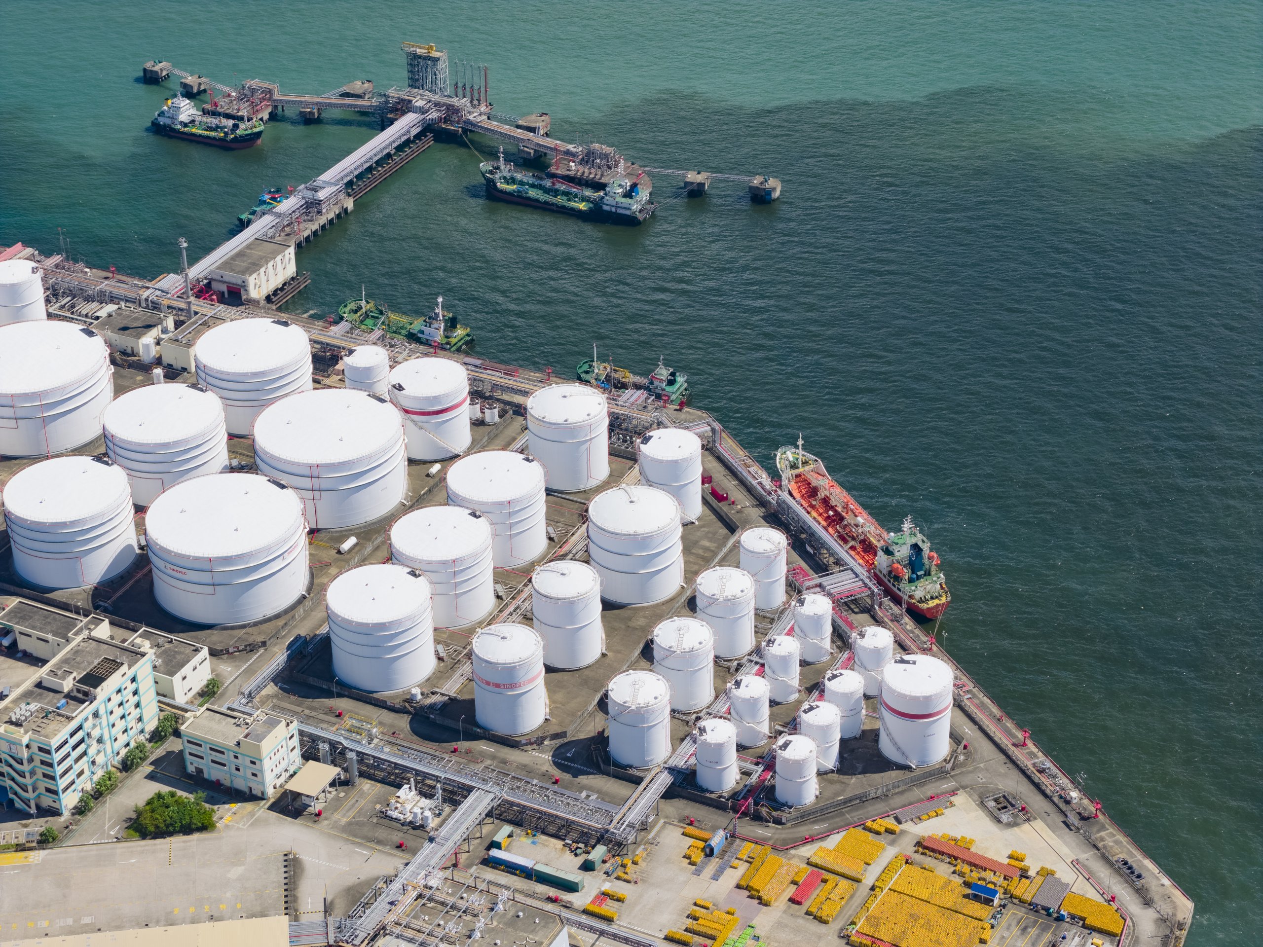 Aerial view of a major petroleum storage terminal with large white tanks, docking piers, and cargo ships in a coastal industrial harbor setting.