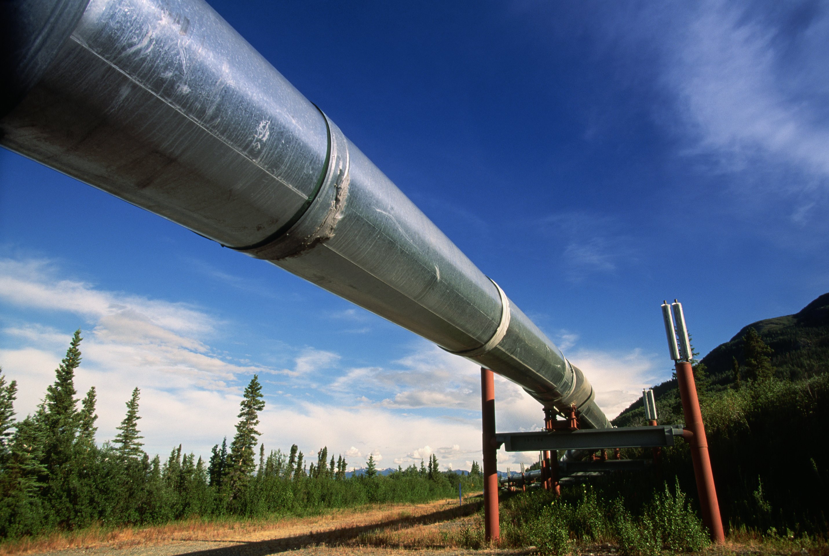 A low-angle shot of a metal pipeline extending through a vast, natural landscape under a bright, expansive blue sky.