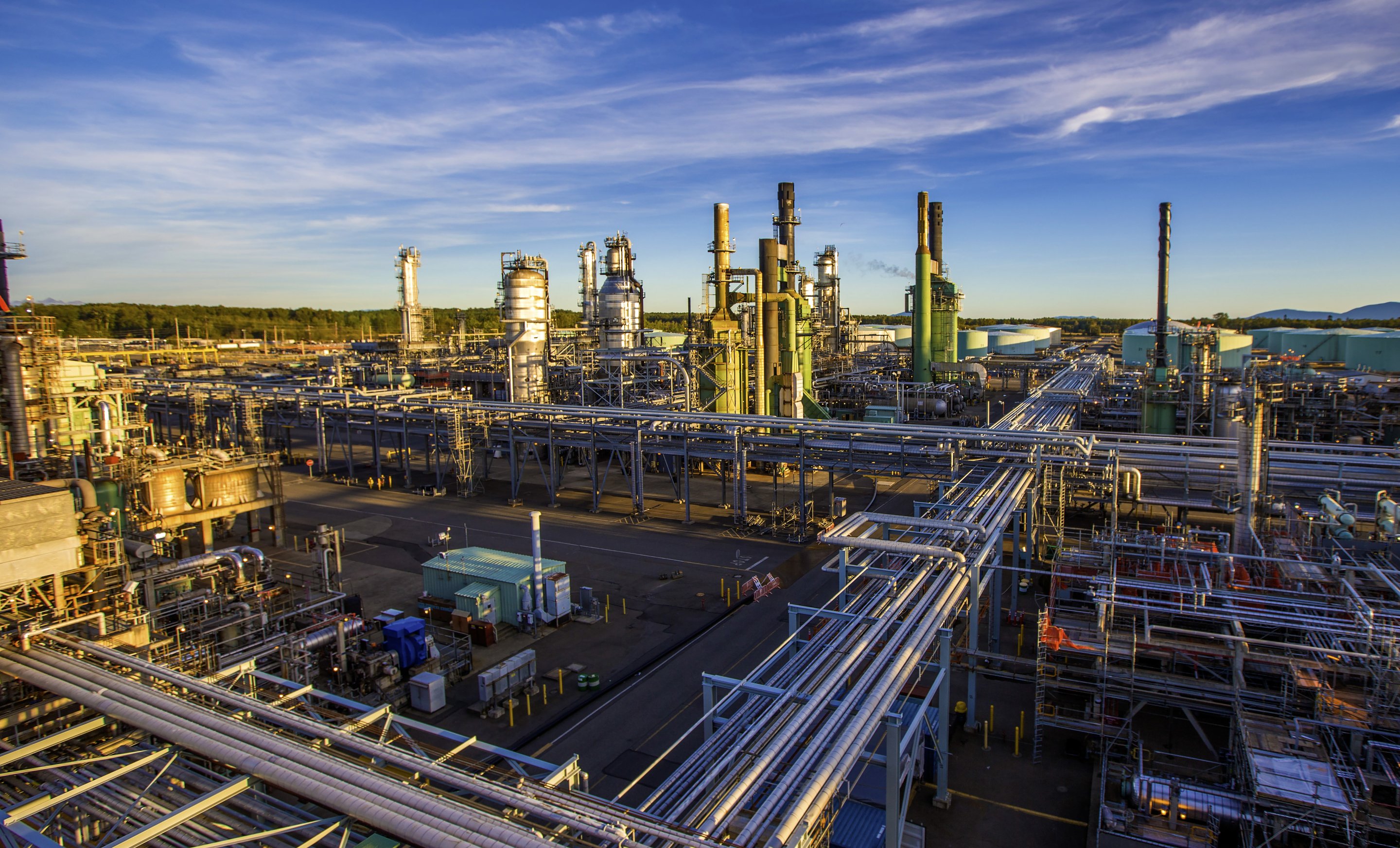 A wide-angle view of a sprawling industrial refinery complex with extensive piping systems and towering processing units under a clear blue sky.