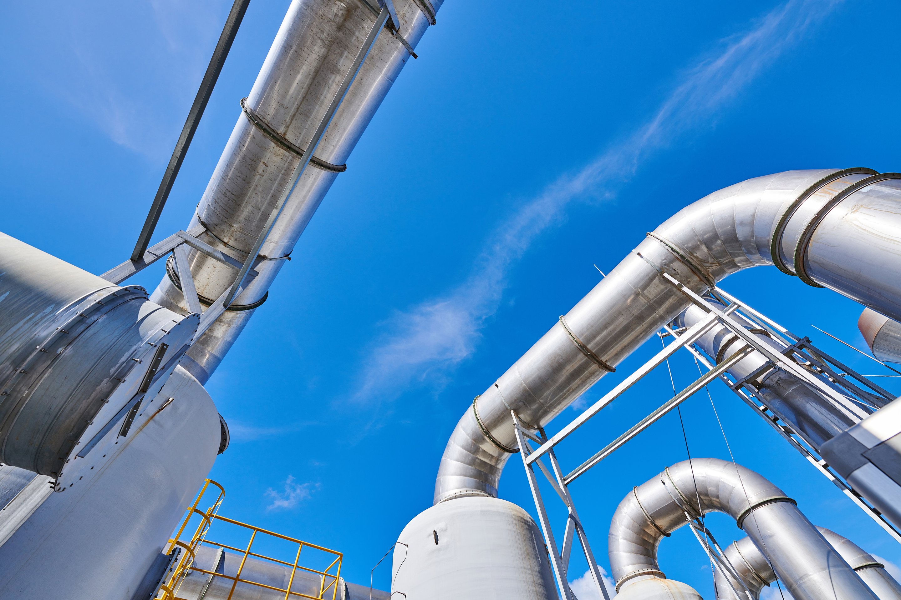 A low-angle view captures complex metal piping and industrial infrastructure against a bright, clear blue sky.