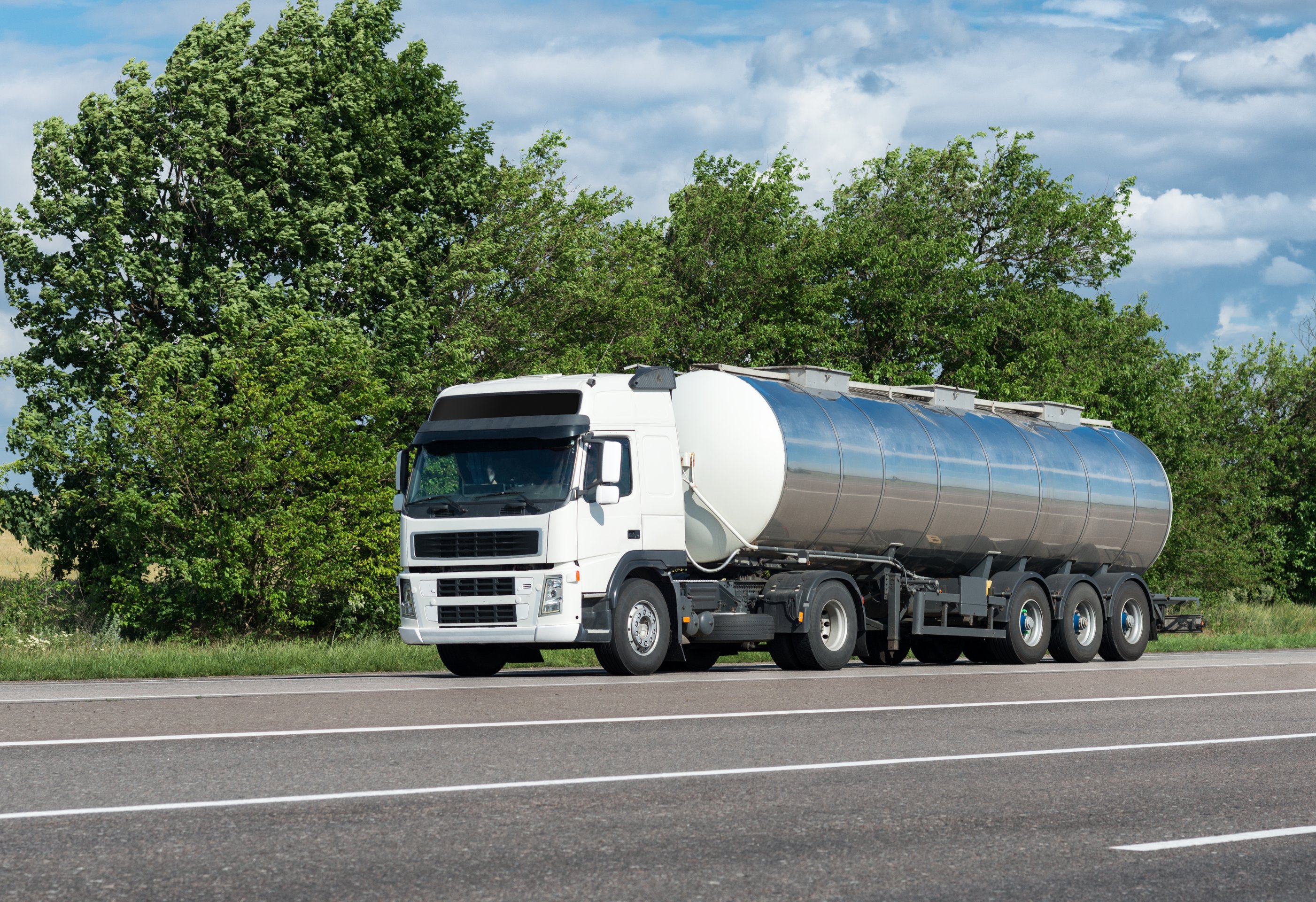 A white semi-truck with a stainless steel tanker trailer drives along a scenic highway lined with lush green trees under a partly cloudy sky.