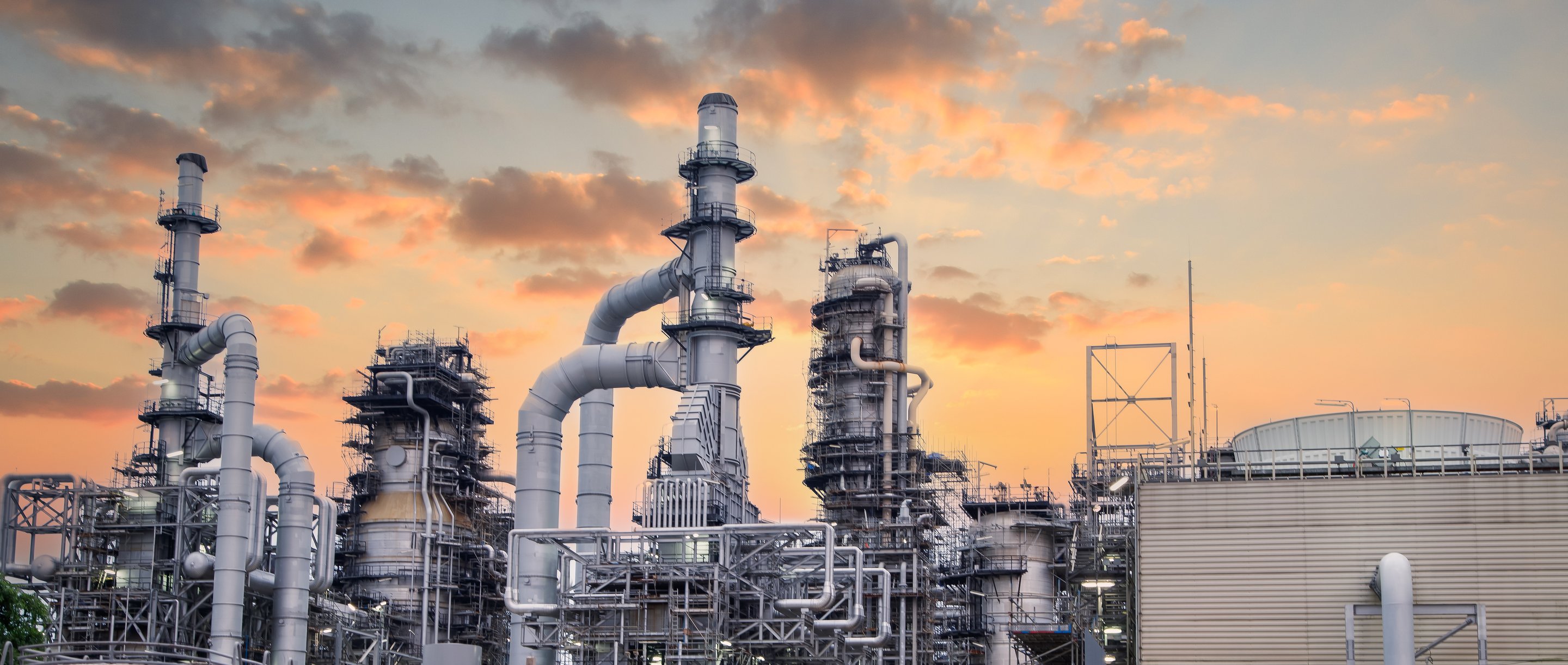 An industrial petrochemical refinery complex silhouetted against a vibrant orange and purple sunset sky.