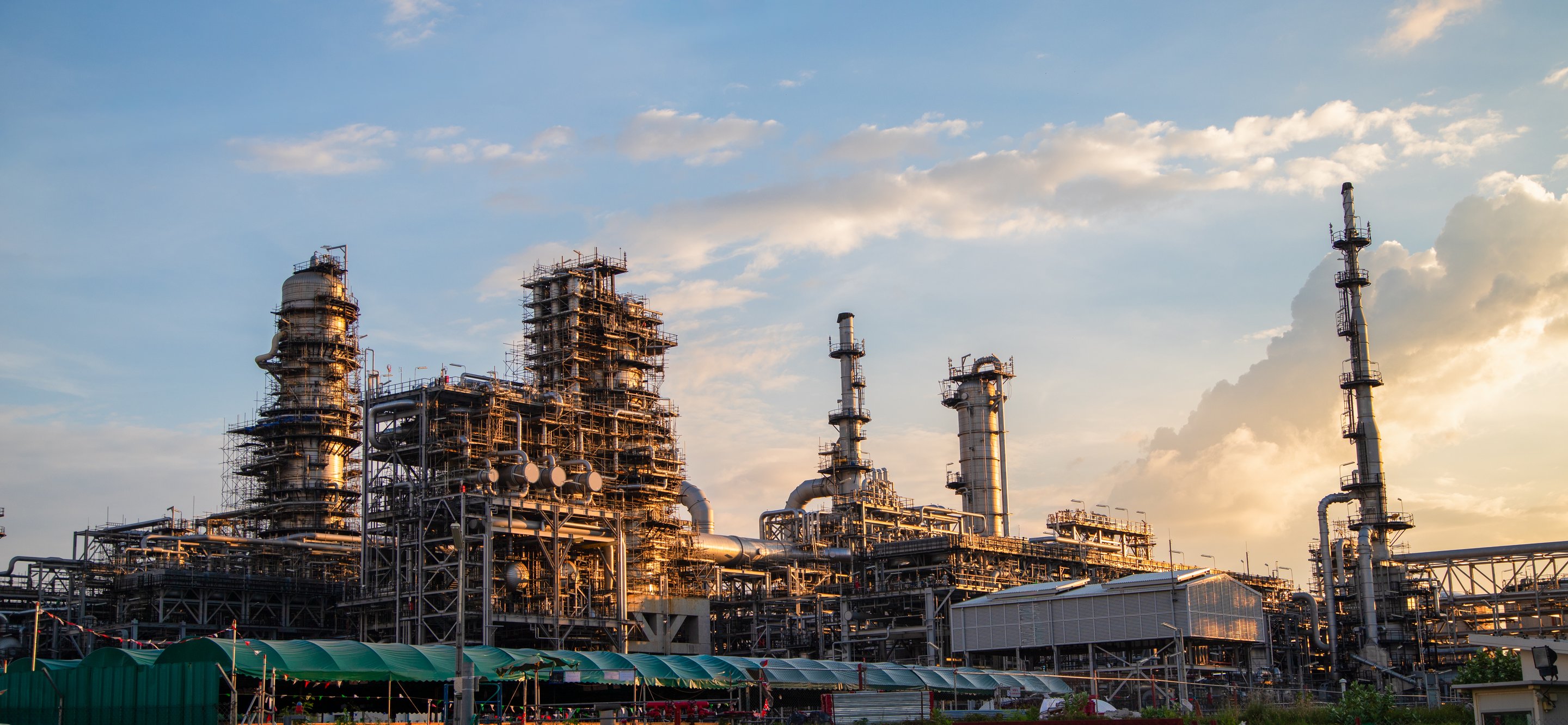 Golden hour illumination highlights the complex piping, scaffolding, and distillation towers of an industrial petrochemical processing plant.