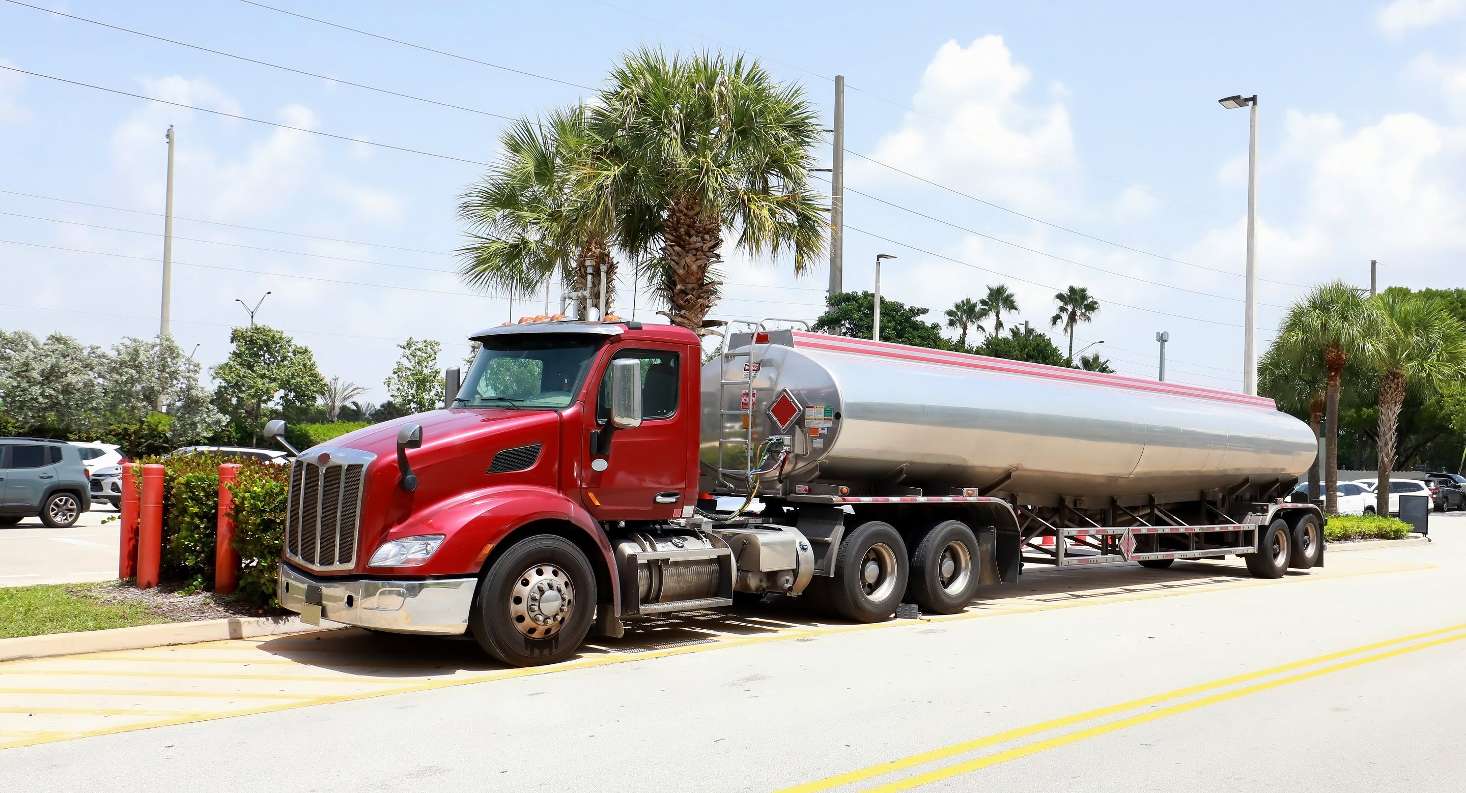 A bright red industrial fuel tanker truck is parked on an asphalt lot near palm trees on a clear, sunny day.