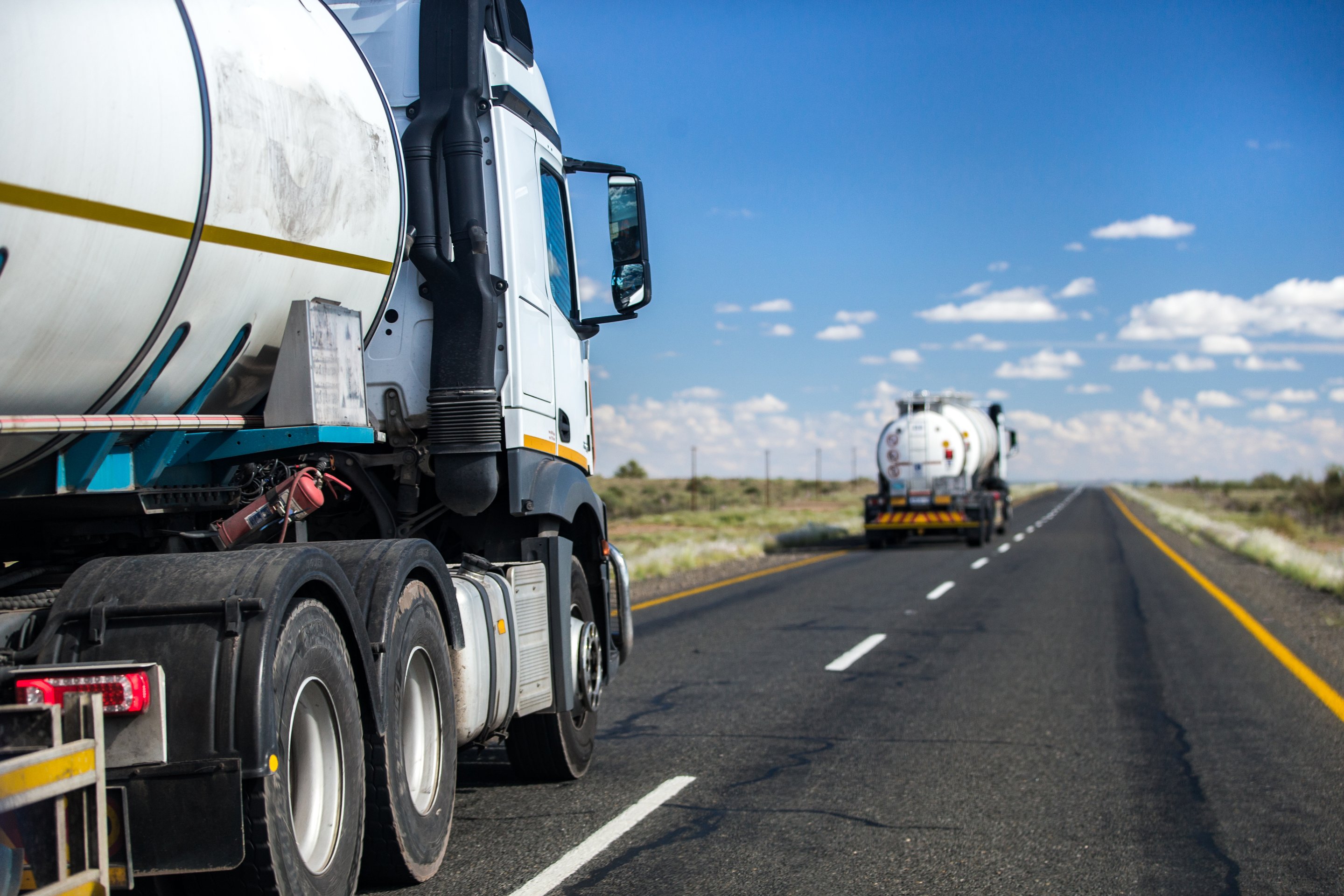 White tanker trucks travel along a long, open highway under a bright blue sky, representing efficient industrial logistics and freight transport.