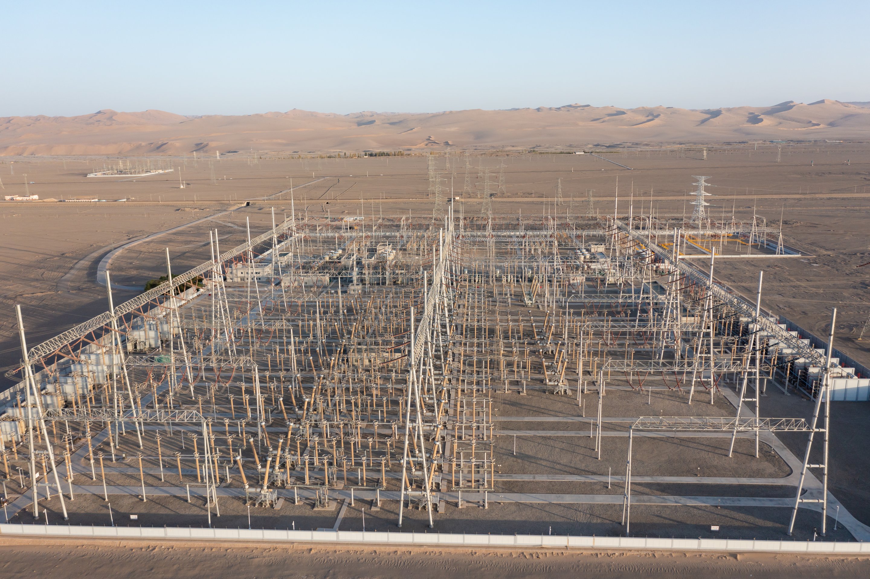 An aerial view captures a large-scale industrial electrical substation situated within a vast, arid desert landscape under a clear sky.