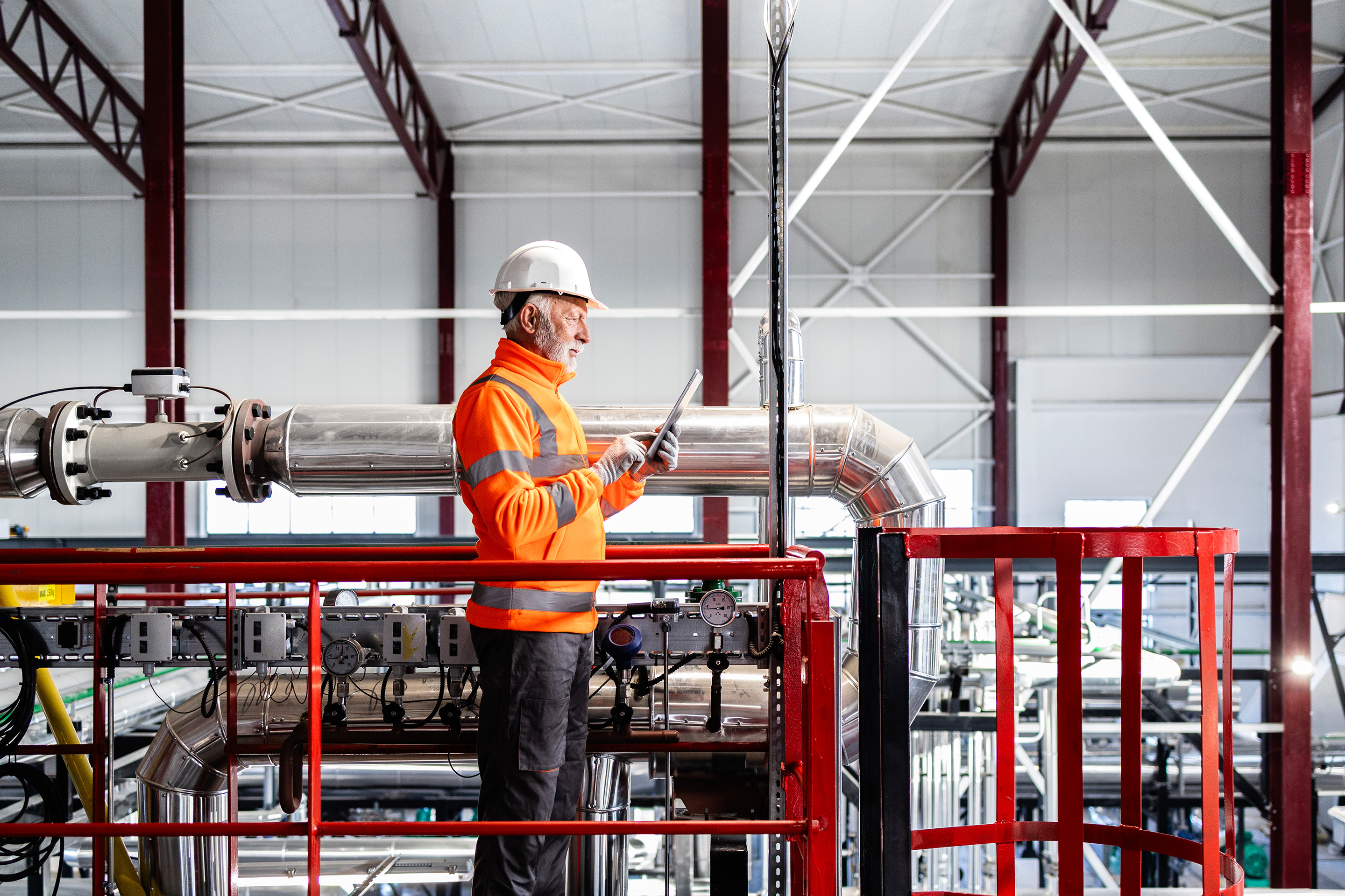 Professional in safety gear reviewing equipment data on a tablet within an industrial plant, emphasising safety compliance, monitoring, and maintenance operations.