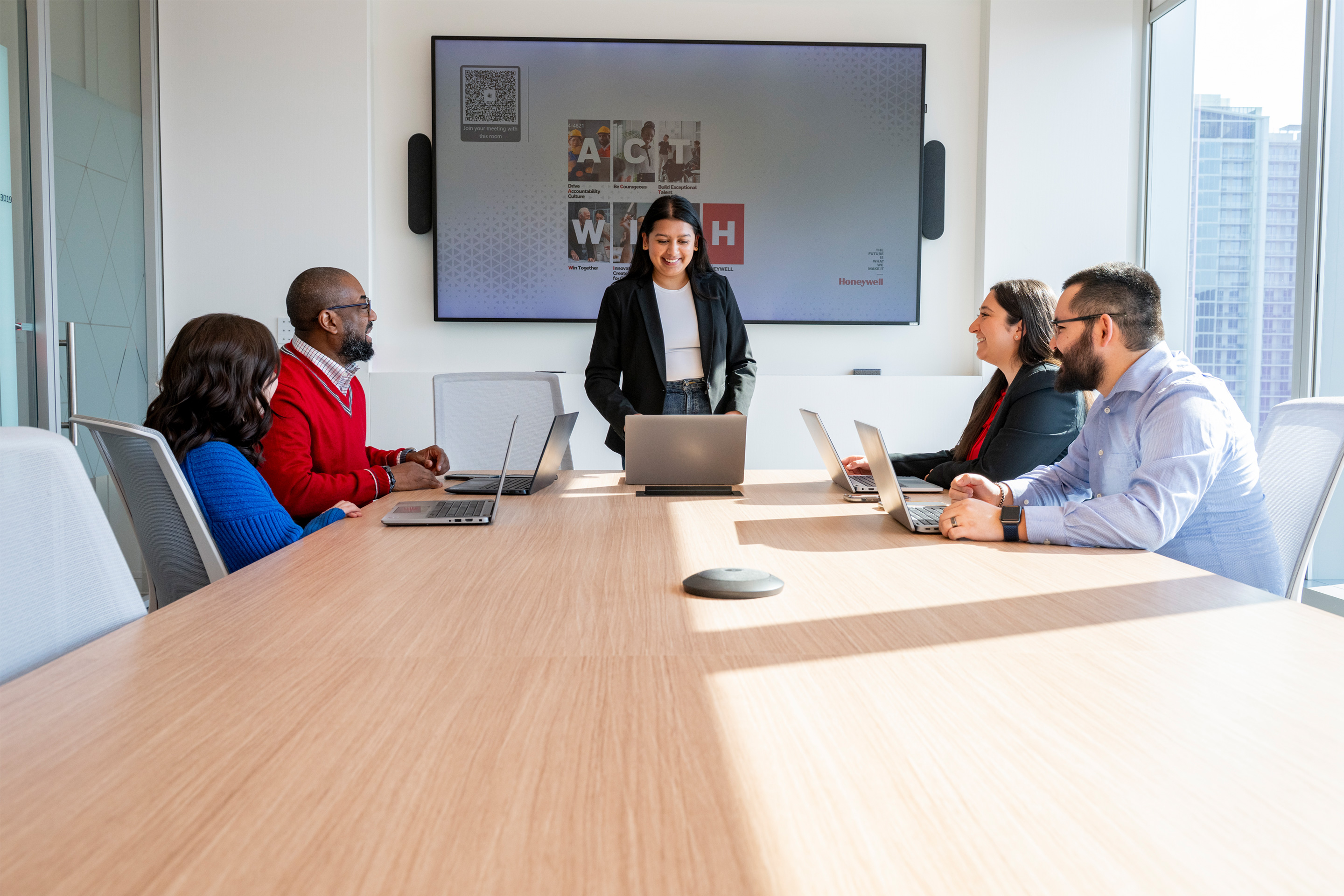 Diverse group of professionals collaborating in a modern office boardroom, with a team lead presenting to colleagues using digital screens, highlighting teamwork, leadership, and corporate culture in a professional workplace environment.