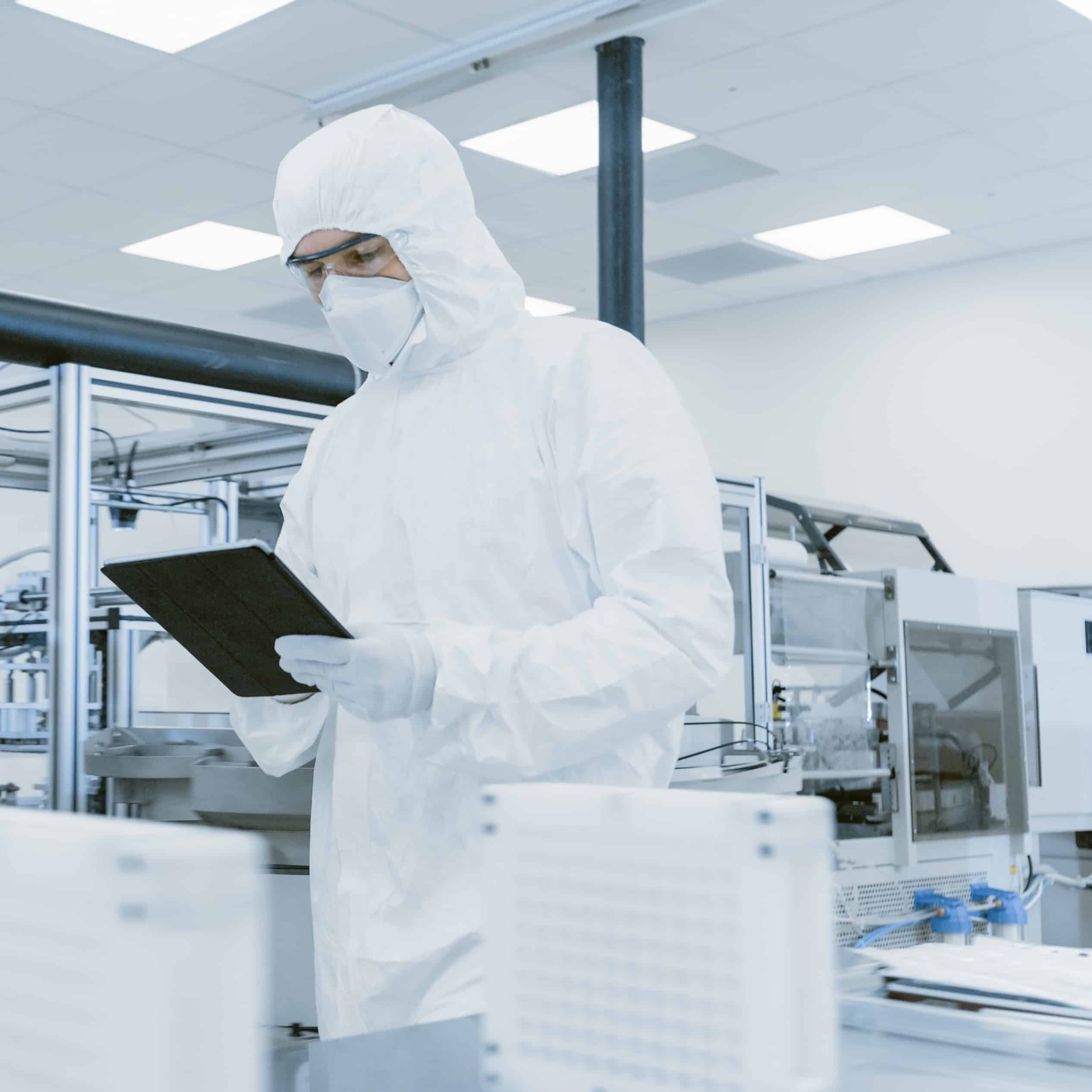 Quality Control Check: Scientist Using Digital Tablet Computer and wearing Protective Suit walks through Manufacturing Laboratory. Product Manufacturing: Pharmaceutics, Semiconductors, Biotechnology.