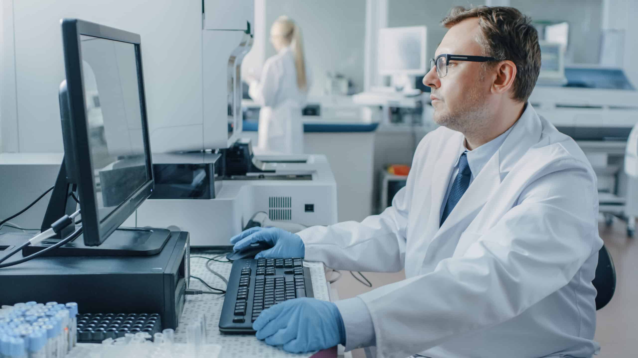 Male Research Scientist Sits at His Workplace in Laboratory, Uses Personal Computer. I the Background Genetics, Pharmaceutical Research Centre with Innovative Equipment.