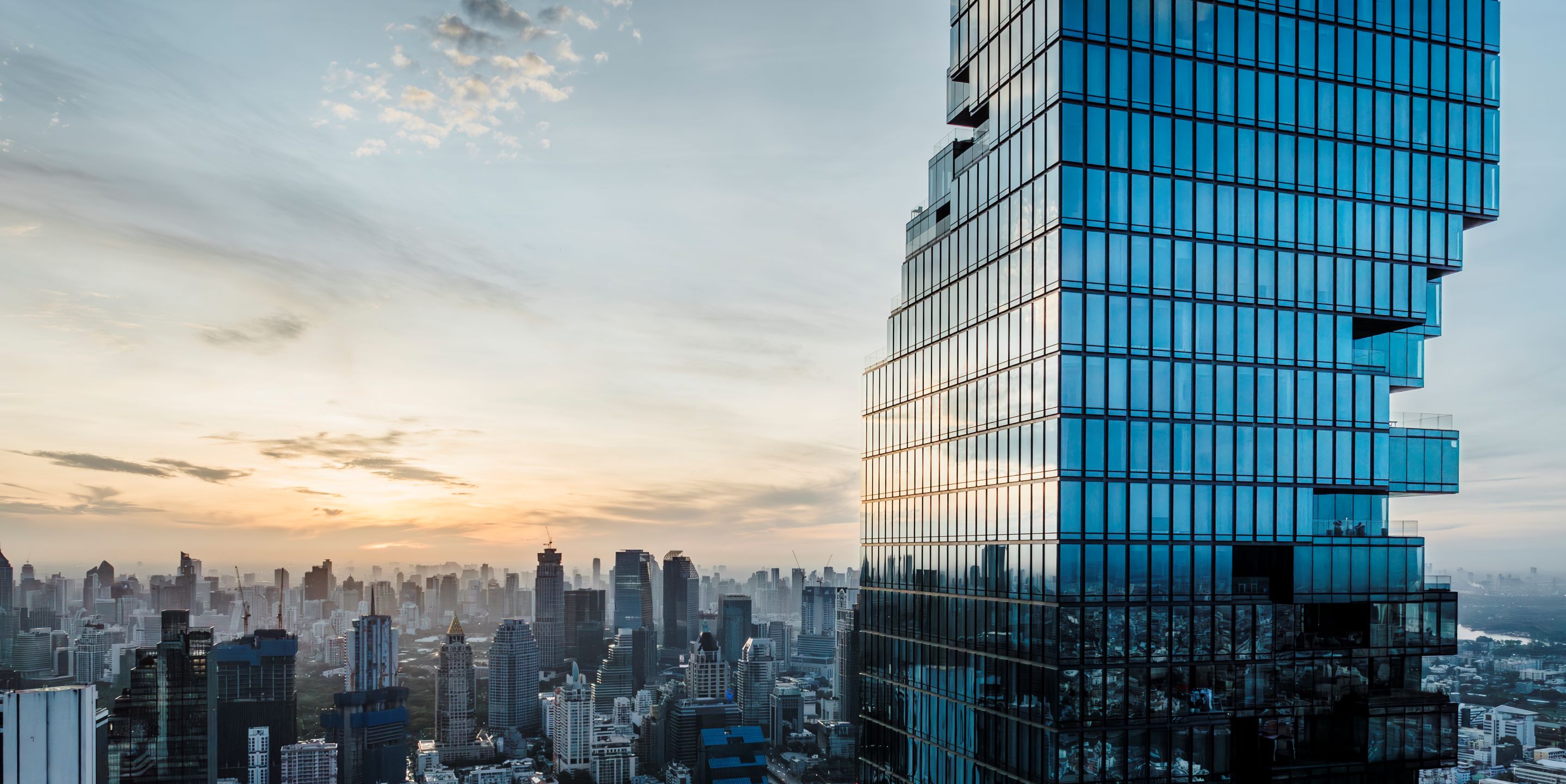 A sleek glass skyscraper stands tall against a sunset backdrop, casting reflections on the cityscape below.