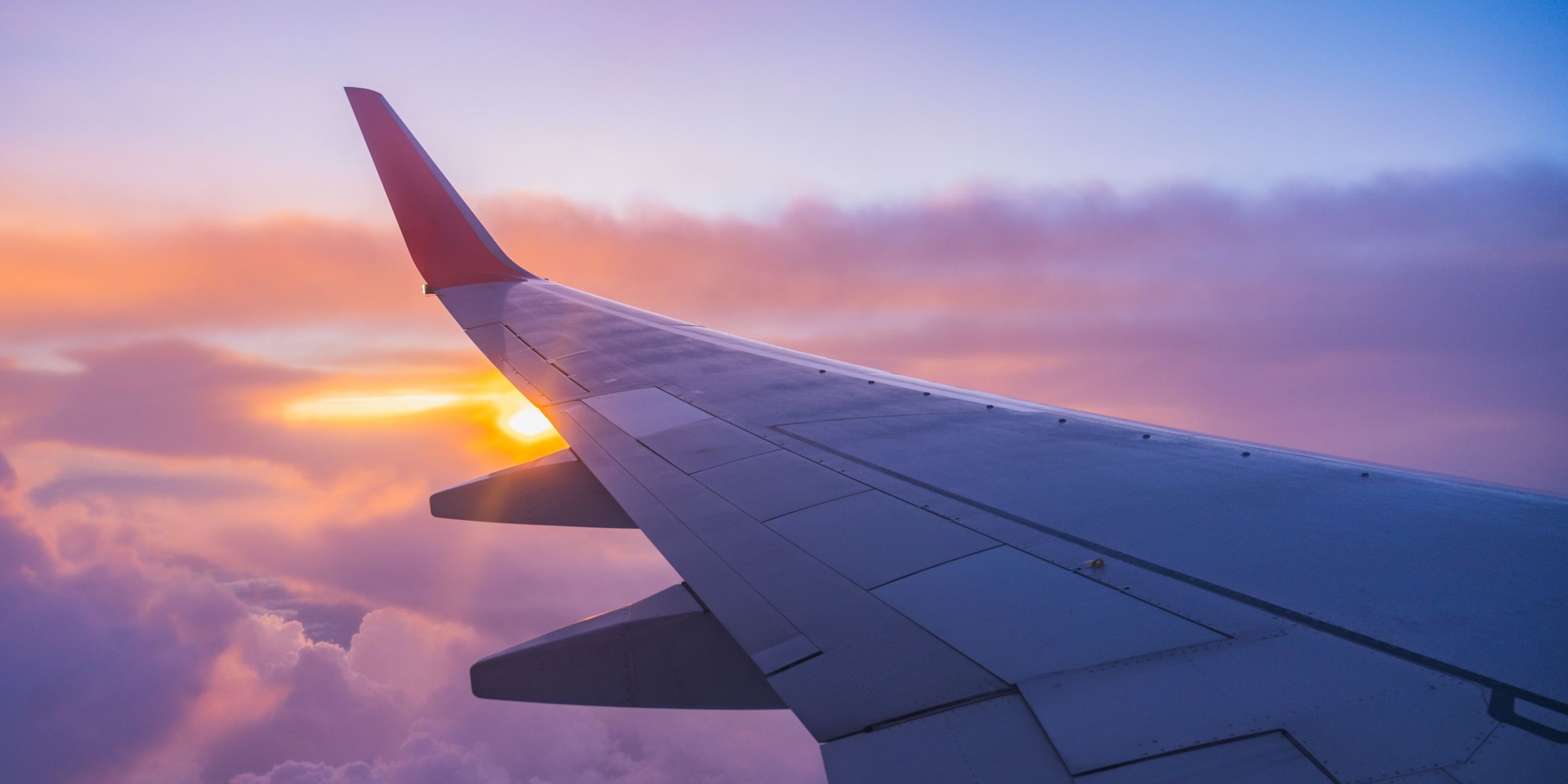 A scenic view from an airplane window showing the aircraft wing silhouetted against a vibrant sky during sunset or sunrise. The gradient of purples and oranges above the clouds captures the beauty of air travel.