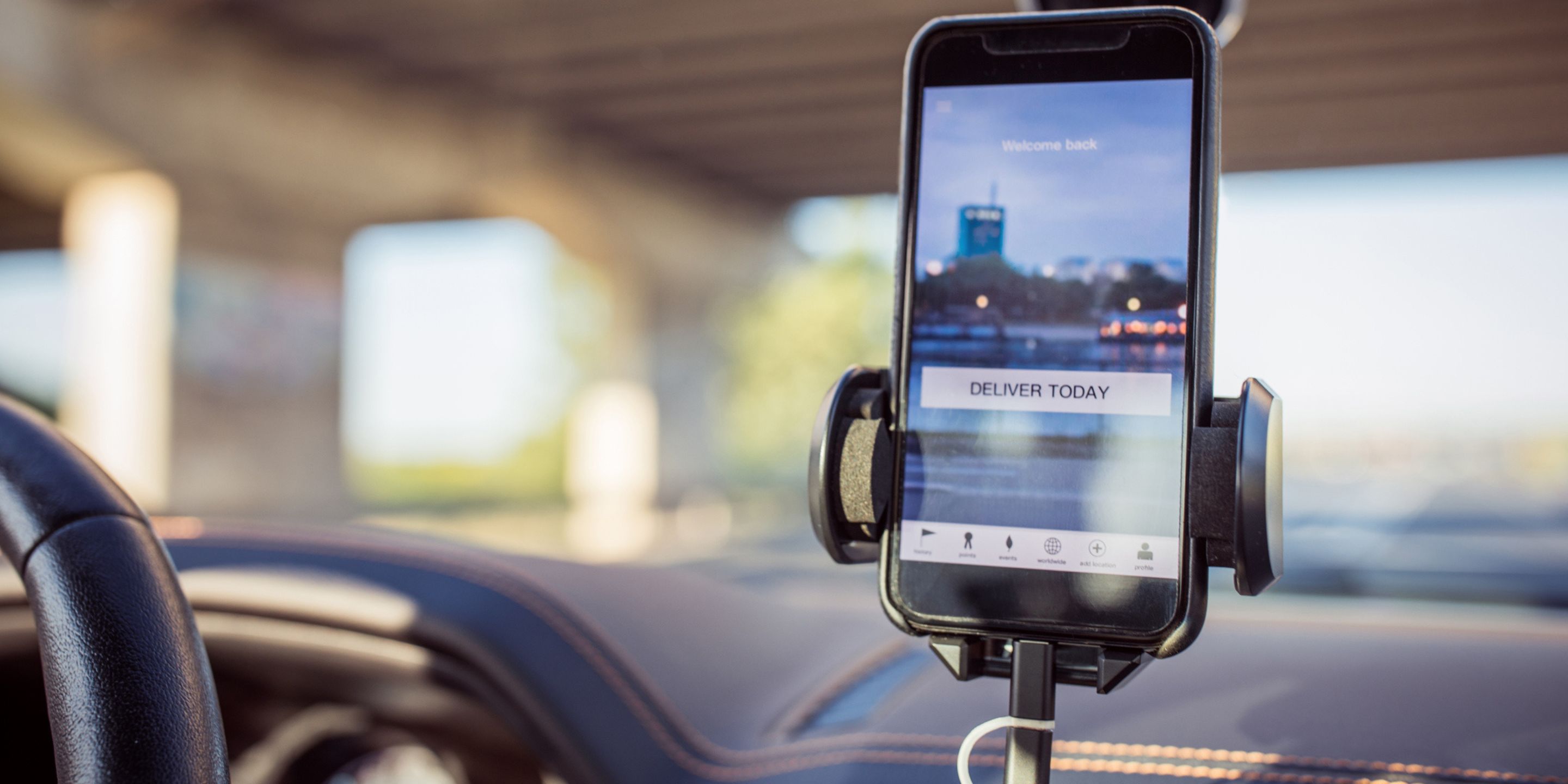 A phone screen shows a city background with the words "Deliver today" in a call-to-action button; the phone is positioned on a mounting device in a car.