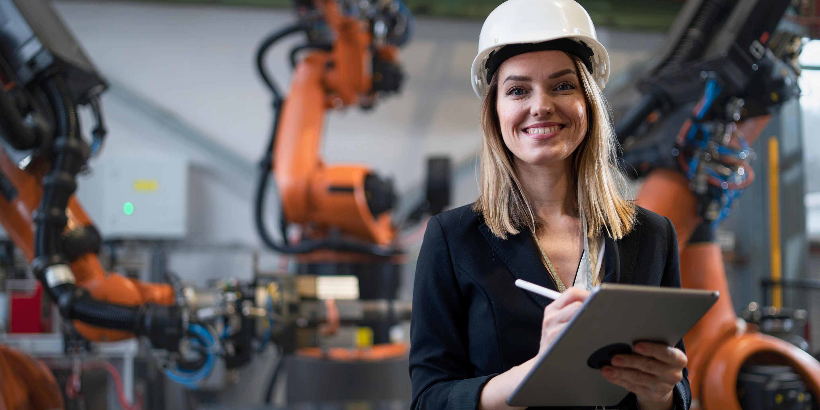 A professional woman wearing a hard hat is working with robotic systems in an industrial environment, showcasing the integration of technology in modern manufacturing.