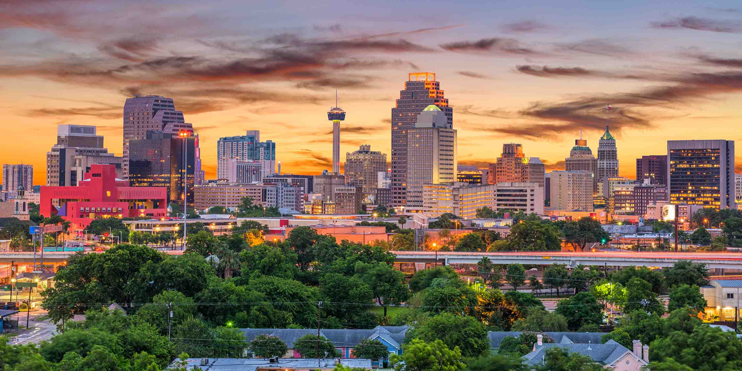 A vibrant sunset casts hues over the San Antonio skyline, featuring modern architecture and the iconic Tower Life Building among lush greenery below.
