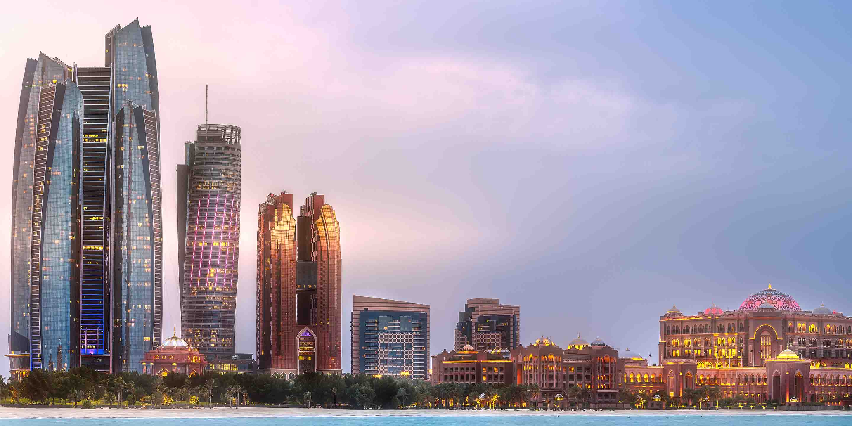 A panoramic view of Abu Dhabi’s skyline at dusk, showcasing modern skyscrapers alongside the ornate Emirates Palace, lit up against a twilight sky.