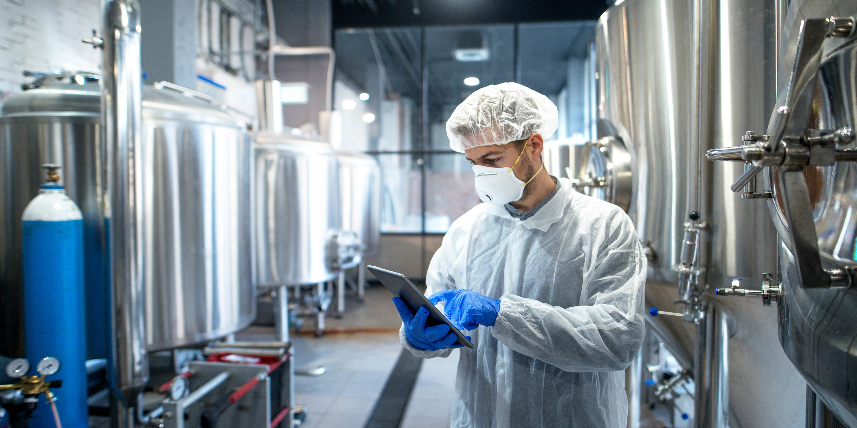 A technician wearing a lab coat and gloves uses a tablet to monitor processes in a modern brewing facility, surrounded by large metal brewing tanks.