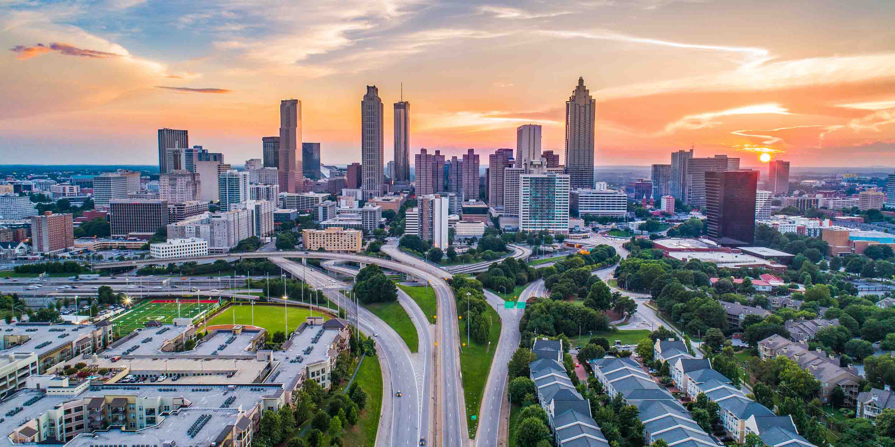 Aerial view of Atlanta’s skyline at sunset, featuring modern skyscrapers and highways amid lush greenery, with a vibrant sky transitioning from blue to orange.