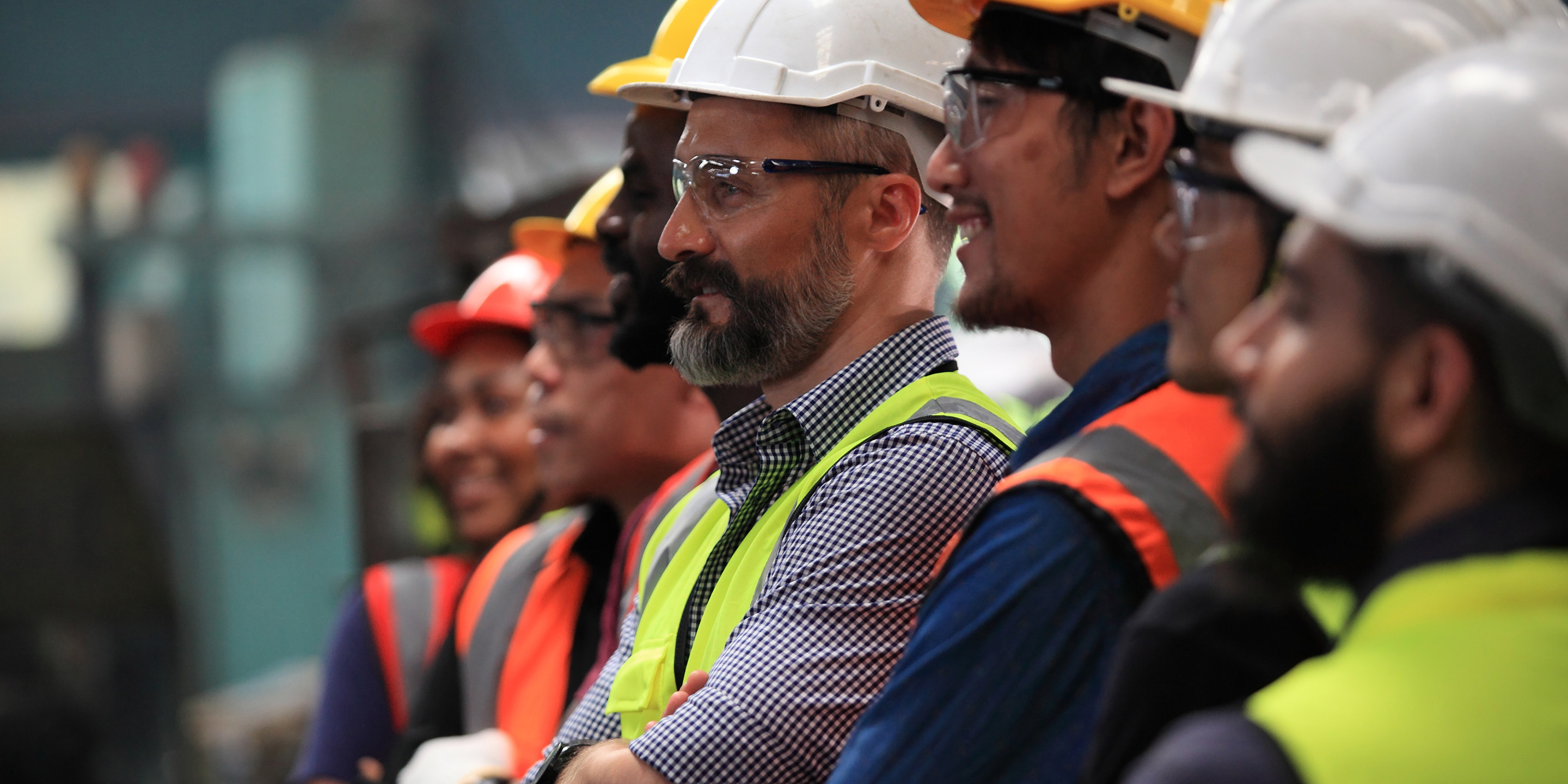 A group of workers in safety helmets and reflective vests, engaged in a discussion in an industrial setting, showcasing teamwork and safety protocols.