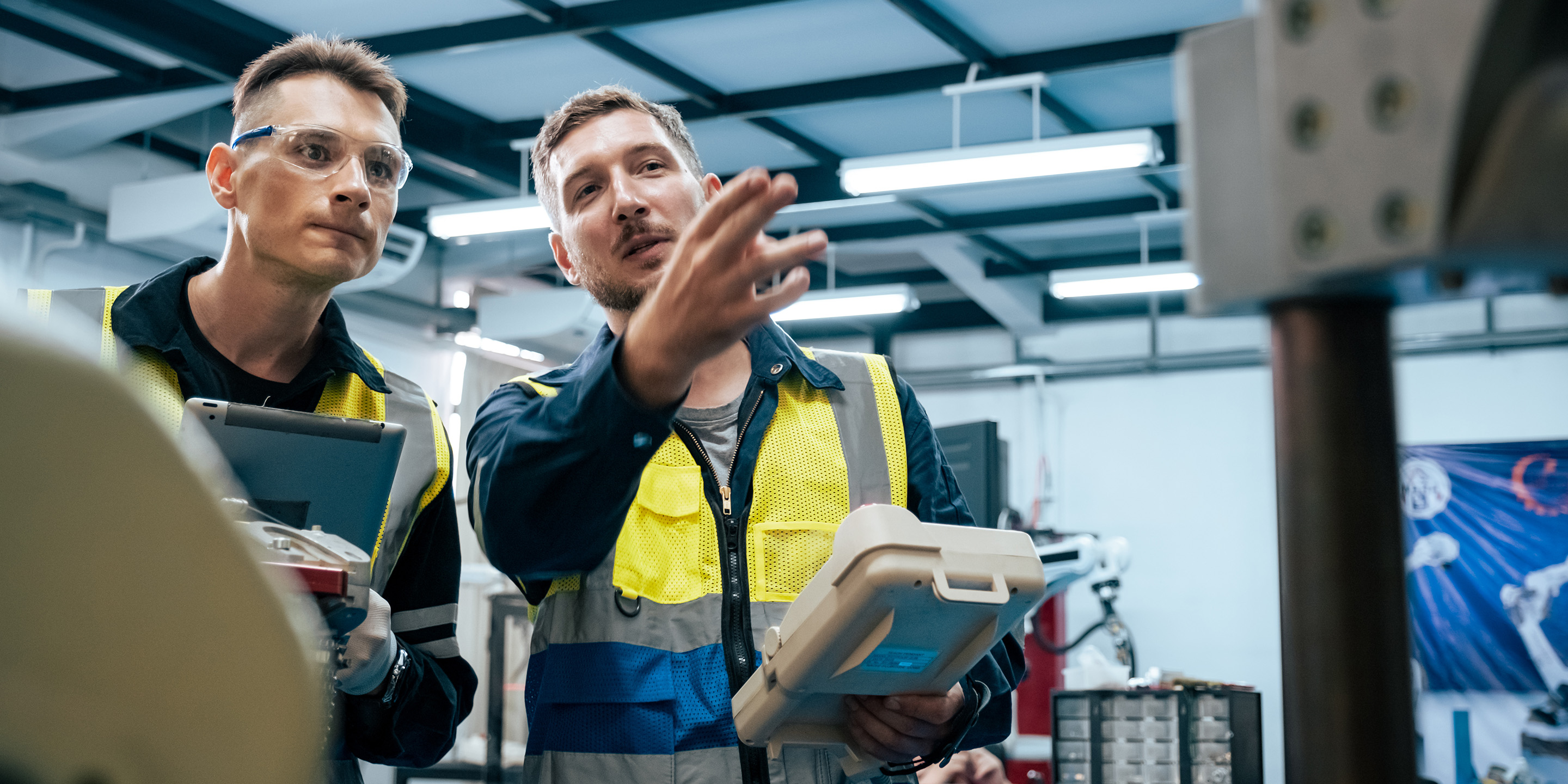 Two engineers in safety vests discuss project details while using a tablet in a well-equipped workshop environment. Tools and machinery are visible in the background.