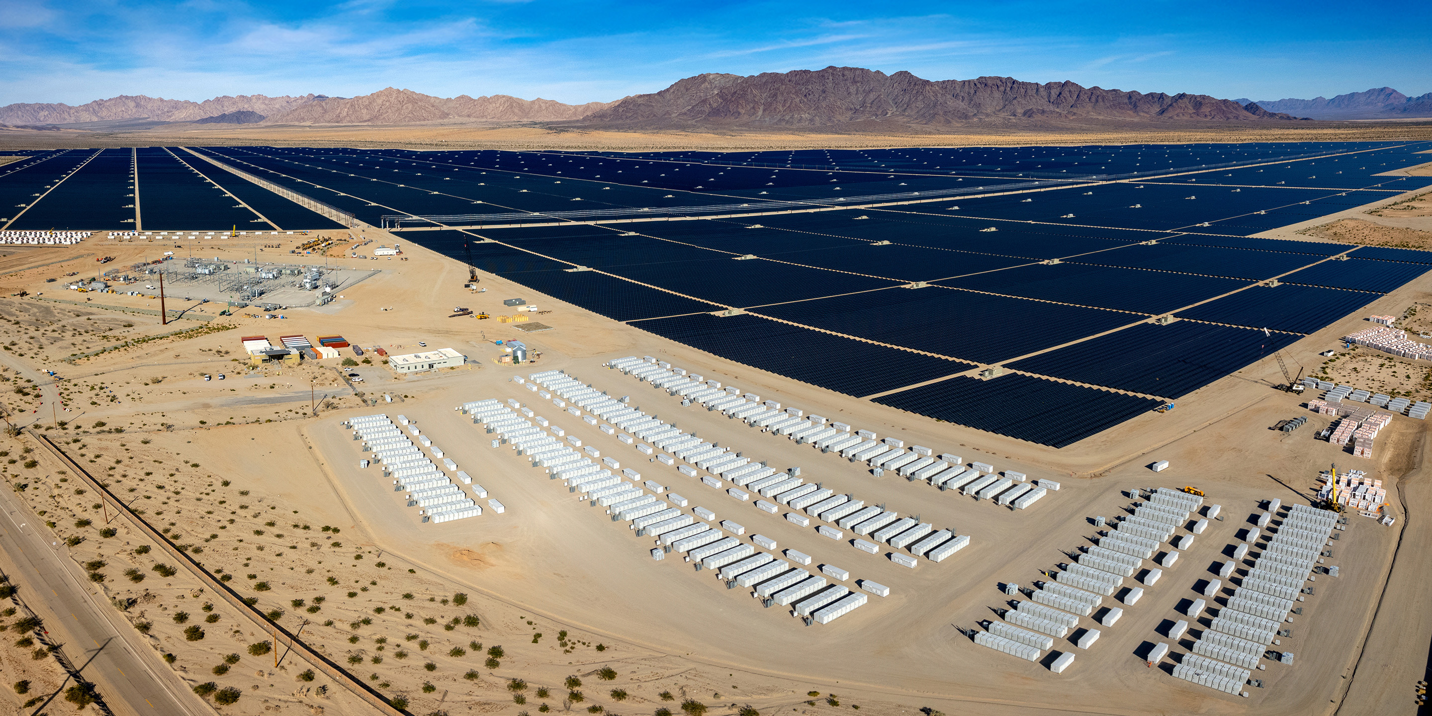 A vast solar power array spreads across desert land, featuring numerous solar panels and energy storage containers, set against a mountainous backdrop.
