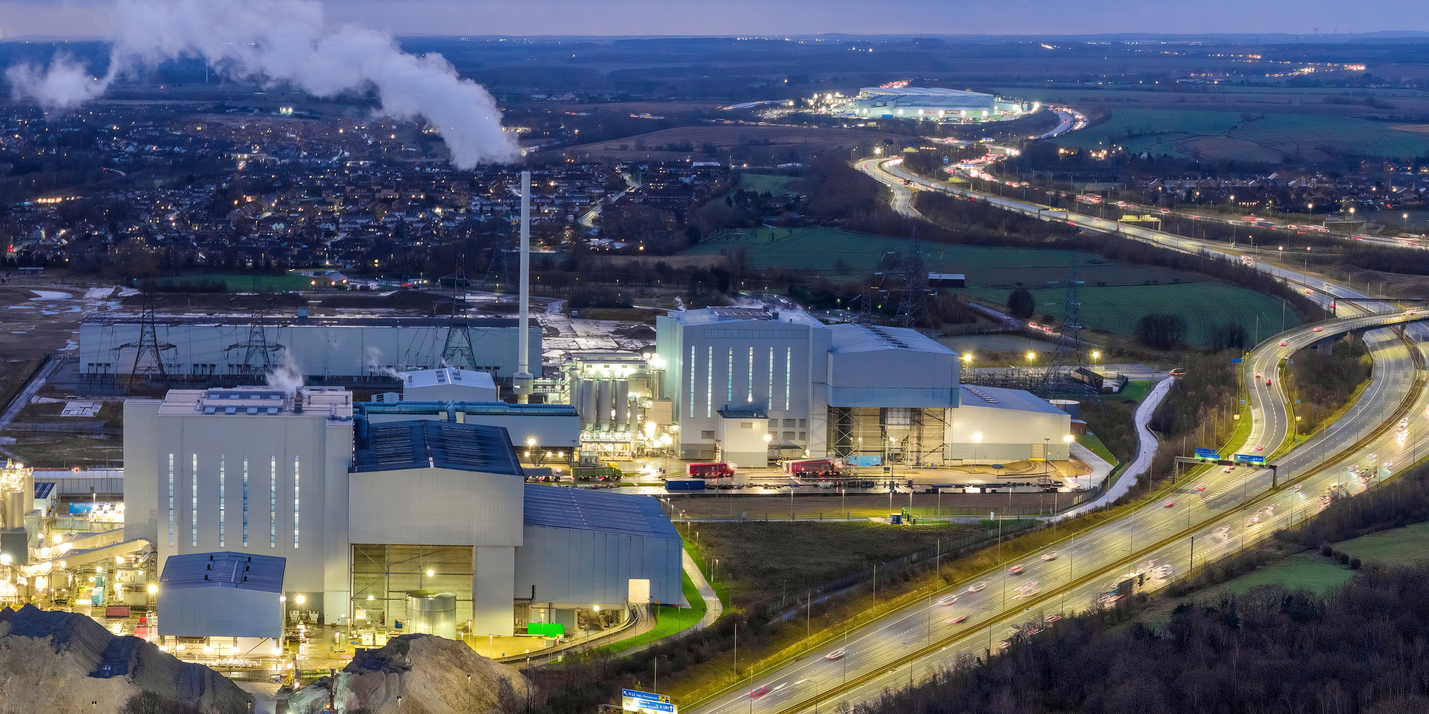 Aerial view of a power plant at dusk, with smoke rising from stacks, adjacent highway traffic, and distant residential areas in a rural landscape.