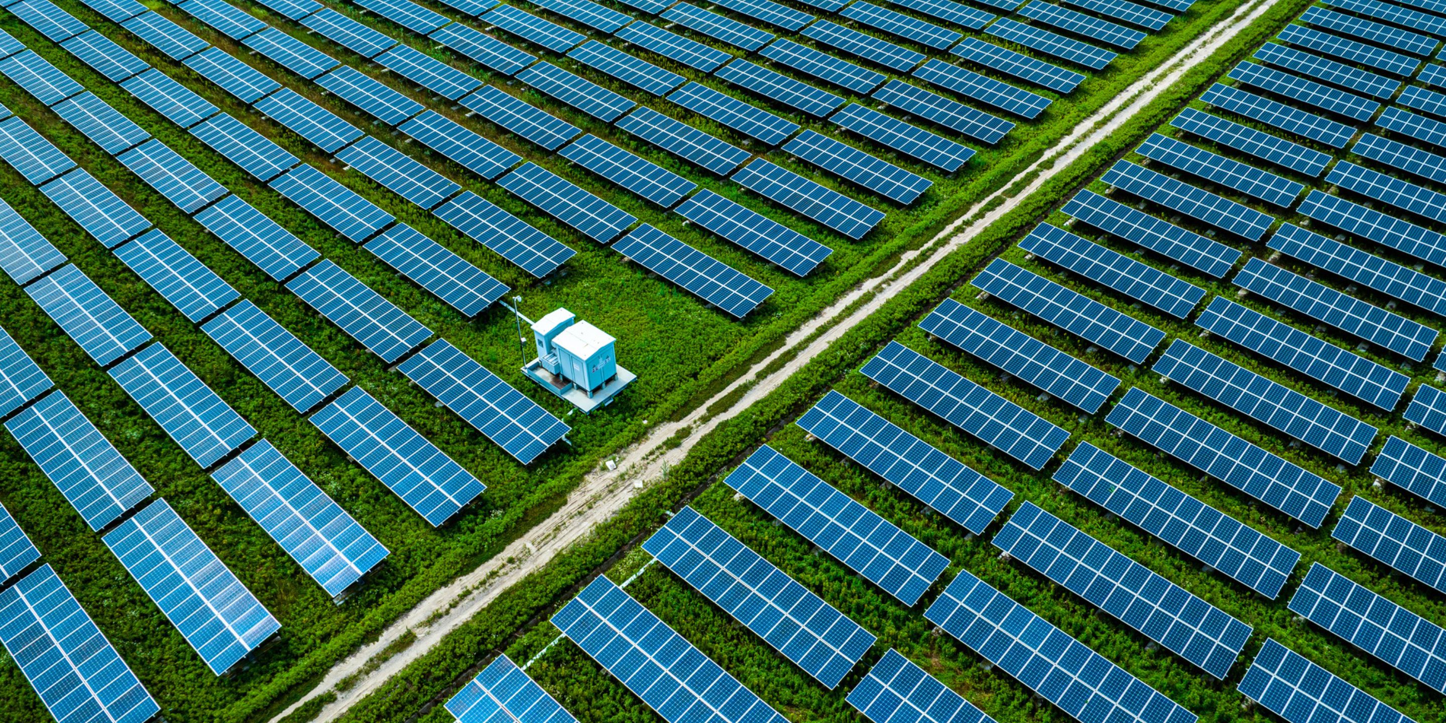 Aerial view of a vast solar panel farm featuring rows of blue solar panels set on green grass. In the foreground, a small blue utility building is visible next to a dirt pathway that runs through the farm, providing access to the organized arrangement of solar panels.
