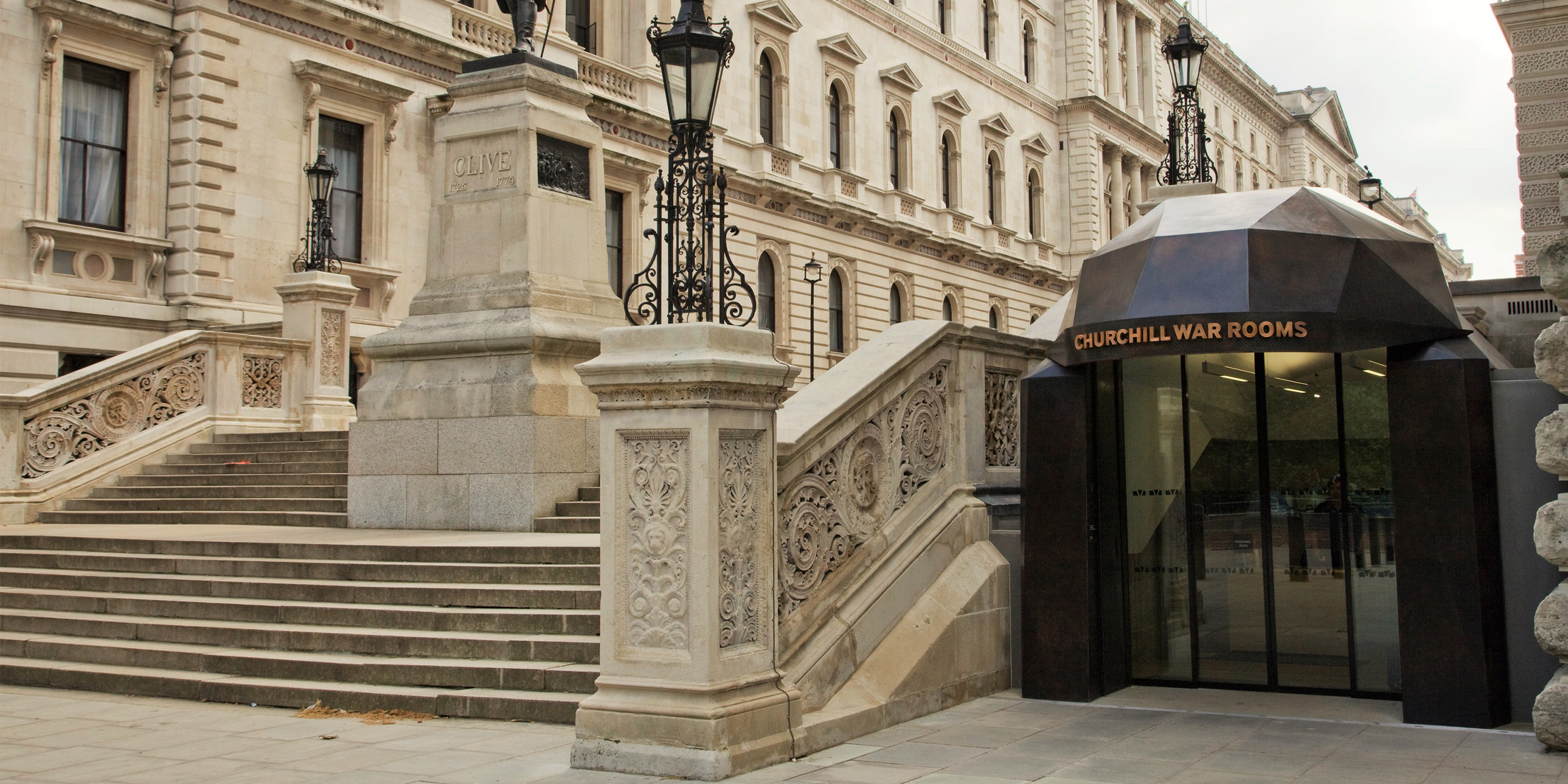 The entrance to the Churchill War Rooms, featuring a modern glass and metal structure with "CHURCHILL WAR ROOMS" inscribed above the door, complemented by ornate stone staircases and historical architecture in the background..