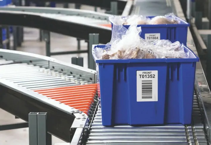An automated conveyor system transports blue plastic bins containing packaged items. Each bin is labeled with barcodes and identifiers, highlighting logistics and inventory tracking in a warehouse or distribution center.