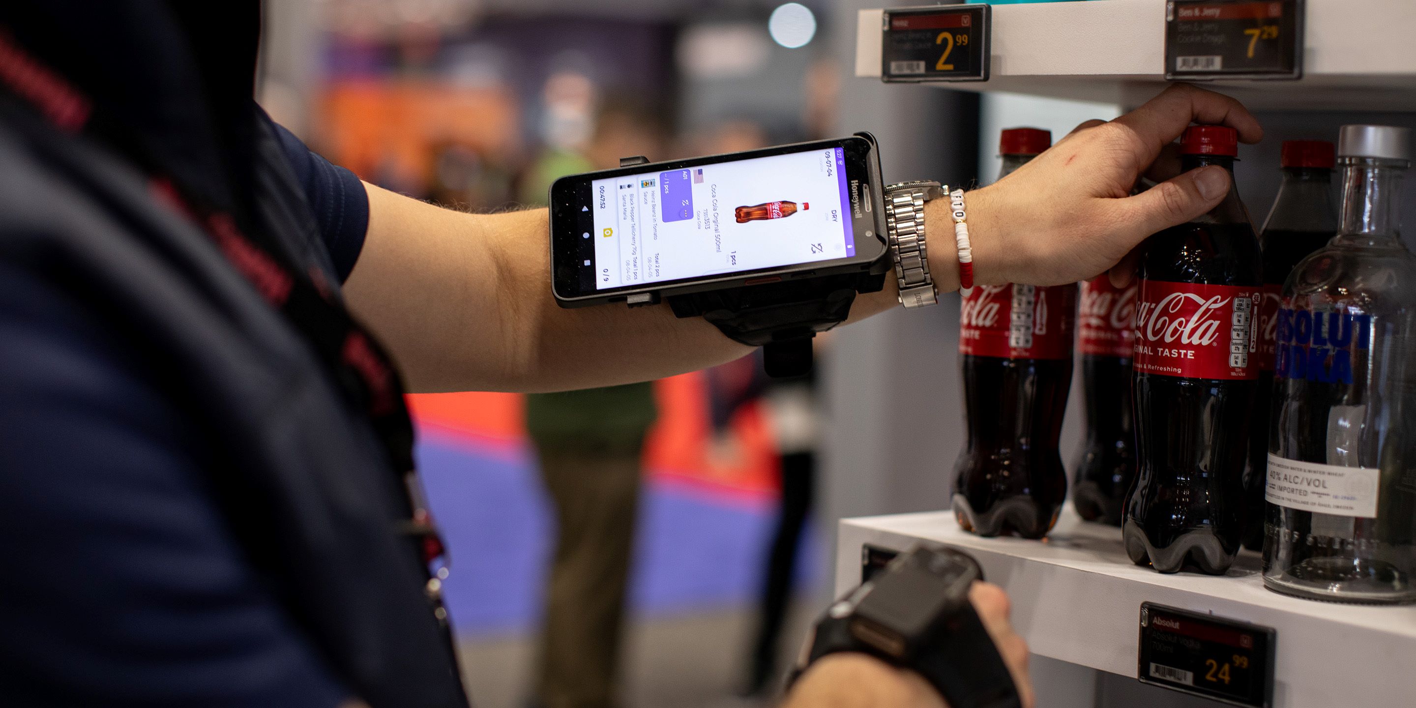 A person using a smartphone mounted on their arm to scan a Coca-Cola bottle from a shelf while managing inventory at a store. The phone screen displays product details and pricing information.