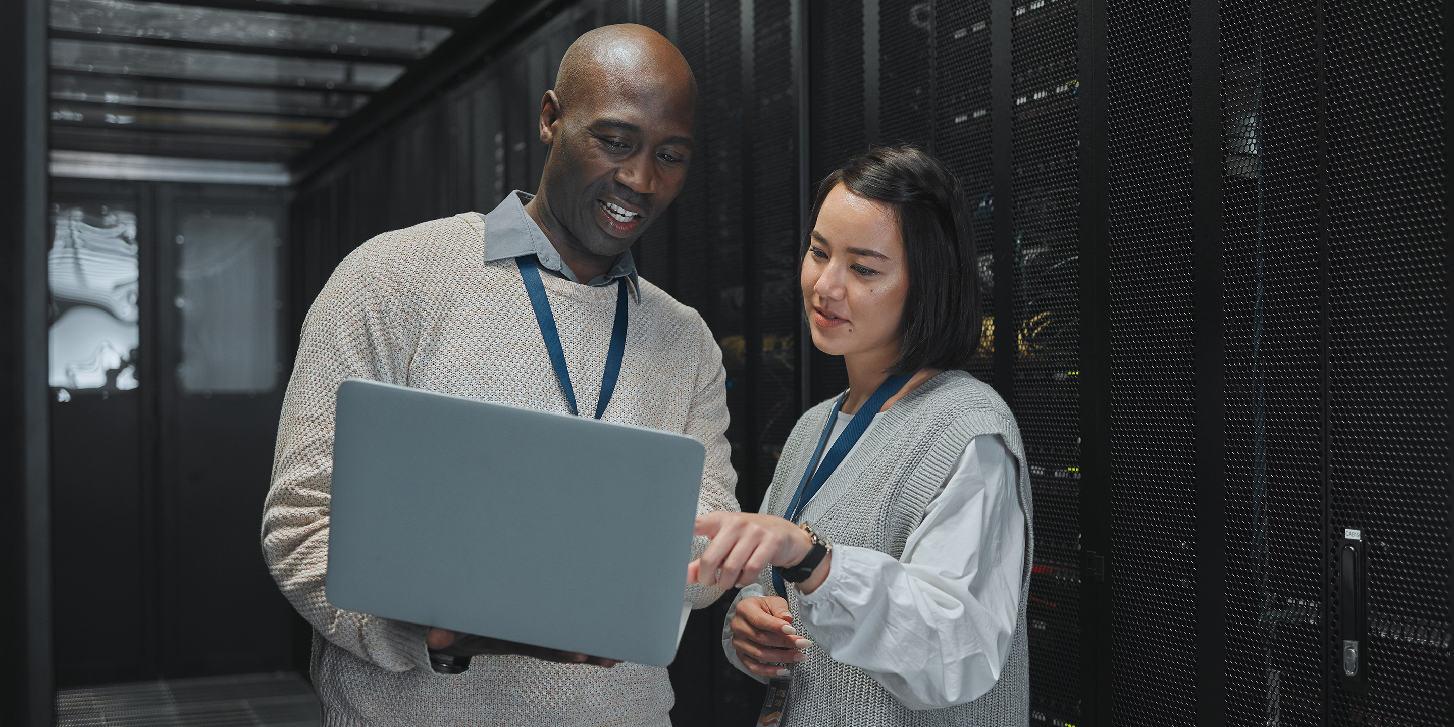 Two professionals in sweaters discussing data analysis while holding a laptop in front of server racks in a data center. Both wear identification lanyards and appear engaged in their conversation.