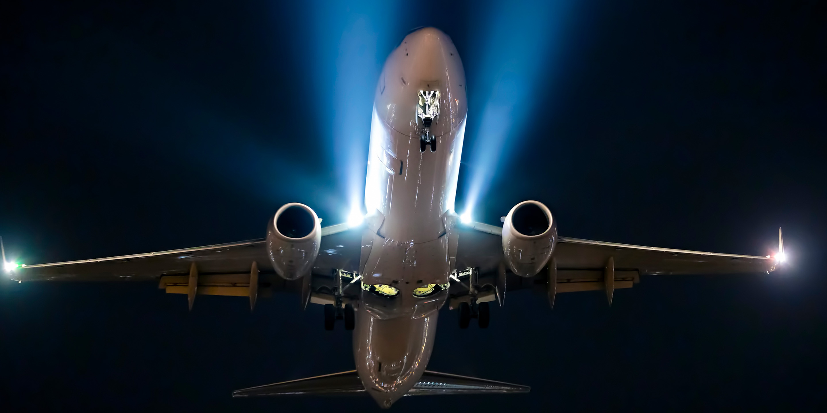 A commercial airplane is shown from below at night, with landing gear extended and bright lights illuminating its undercarriage and engines. The dark sky contrasts with the aircraft’s lighting, highlighting its structure and motion.