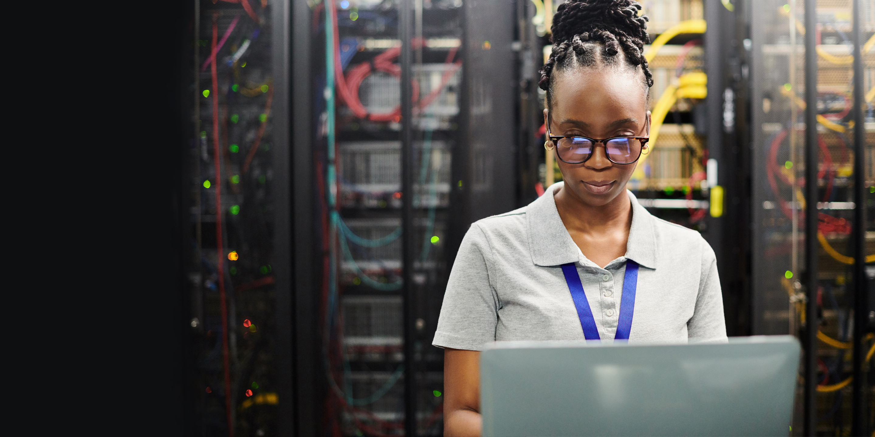 An IT professional stands in a data center holding an open laptop, facing server racks filled with cables and indicator lights. The image highlights infrastructure management, system monitoring, and enterprise IT operations.